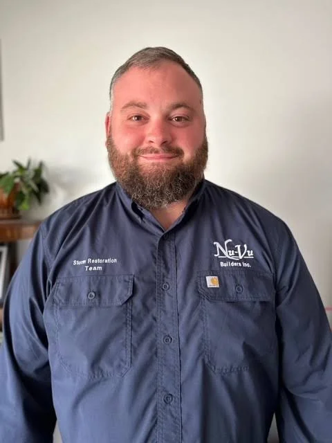 A man with a beard smiling, wearing a navy blue Carhartt shirt with company logos, standing in front of a white wall with a small plant on a table to his left.