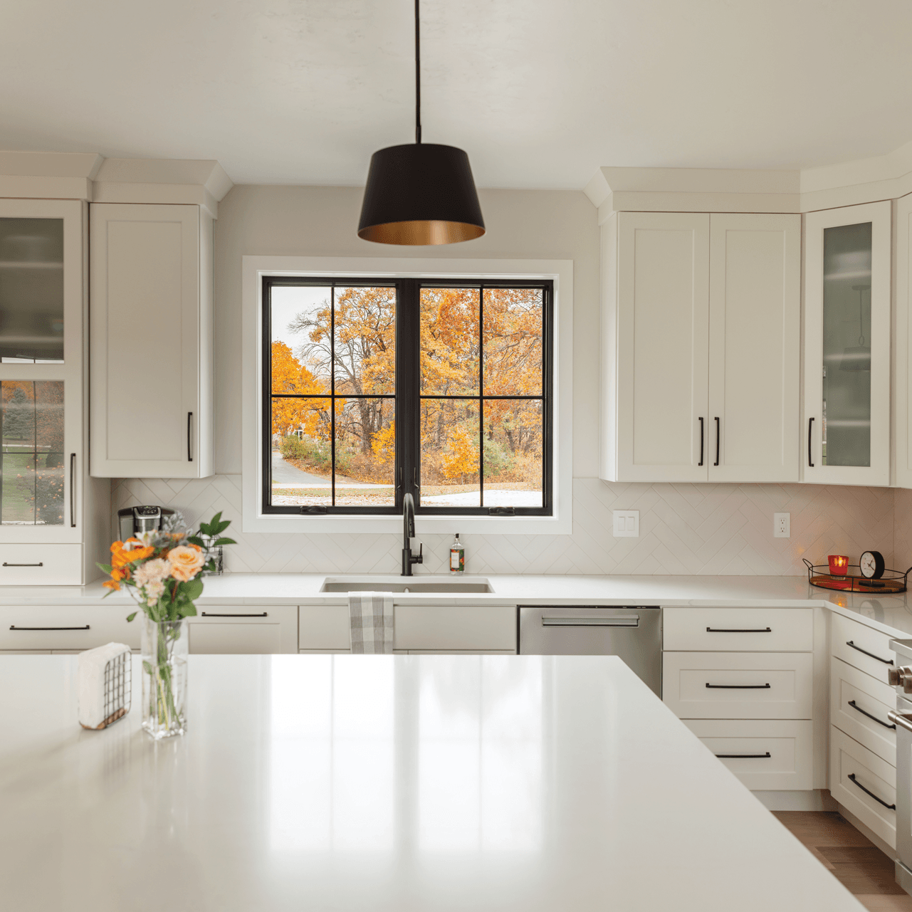 Modern kitchen with white cabinets, a black window with outside autumn trees, and a white countertop with a vase of flowers.