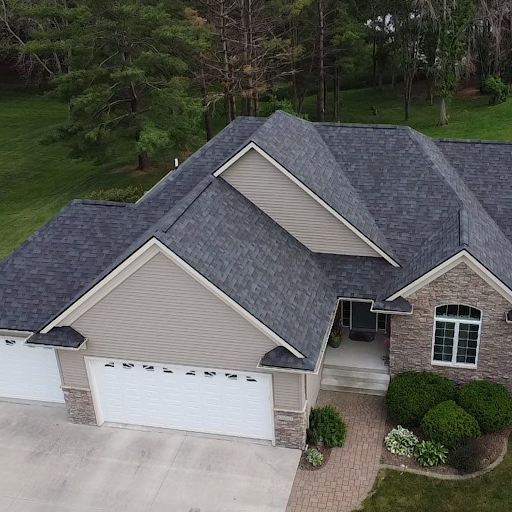 Aerial view of a suburban house with a multi-gabled dark gray shingled roof, beige siding, and a white garage door. The house is surrounded by a concrete driveway, a brick pathway leading to the front porch, and well-maintained bushes and flower beds