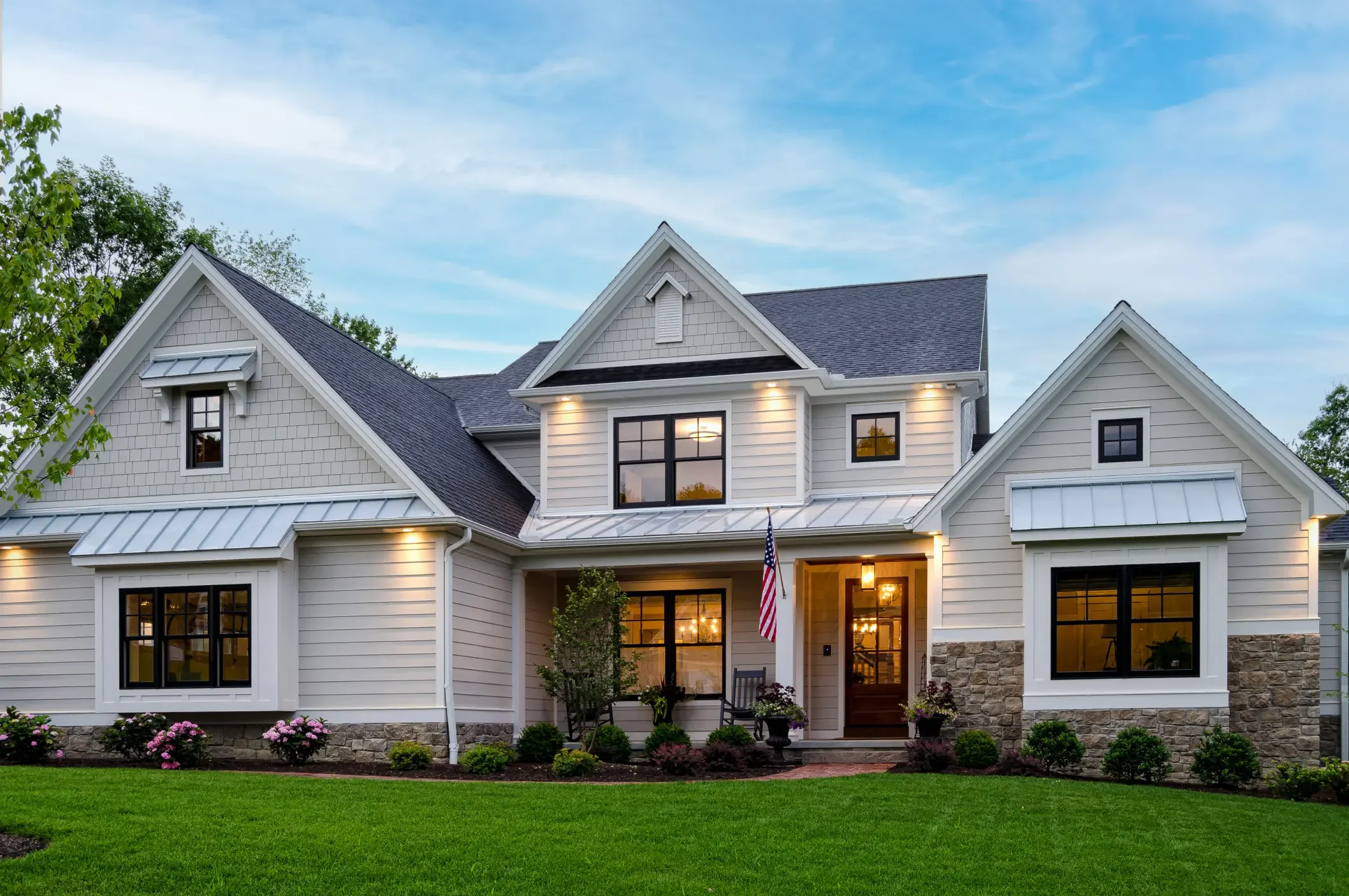 A two-story white house with black window frames, a gabled roof, and a porch is surrounded by green grass and flowering bushes. The house has several windows, some with shutters, and exterior lighting illuminating the facade. An American flag hangs n