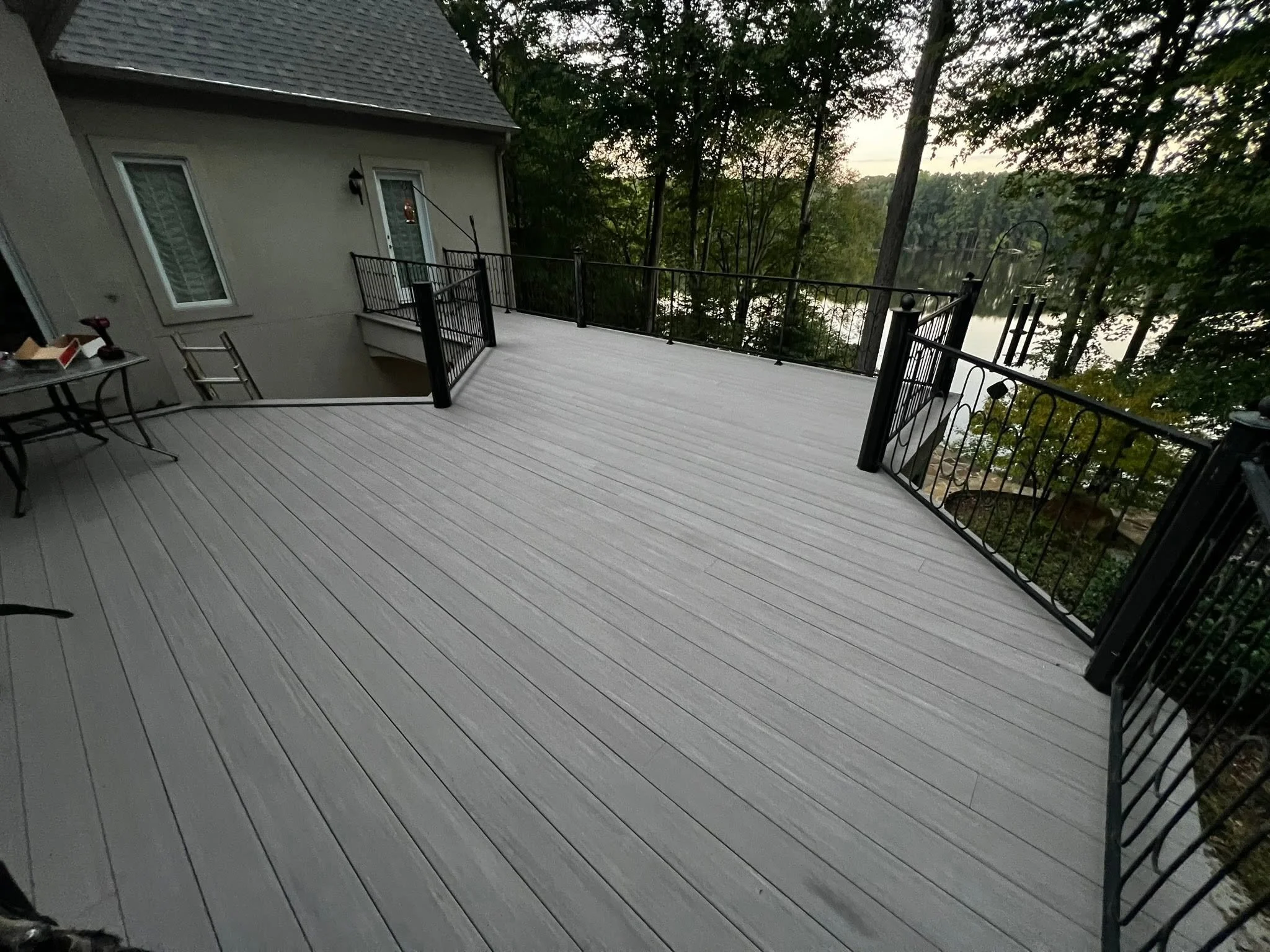 A spacious elevated wooden deck with black metal railings, overlooking a calm lake surrounded by trees in a natural setting at dusk.