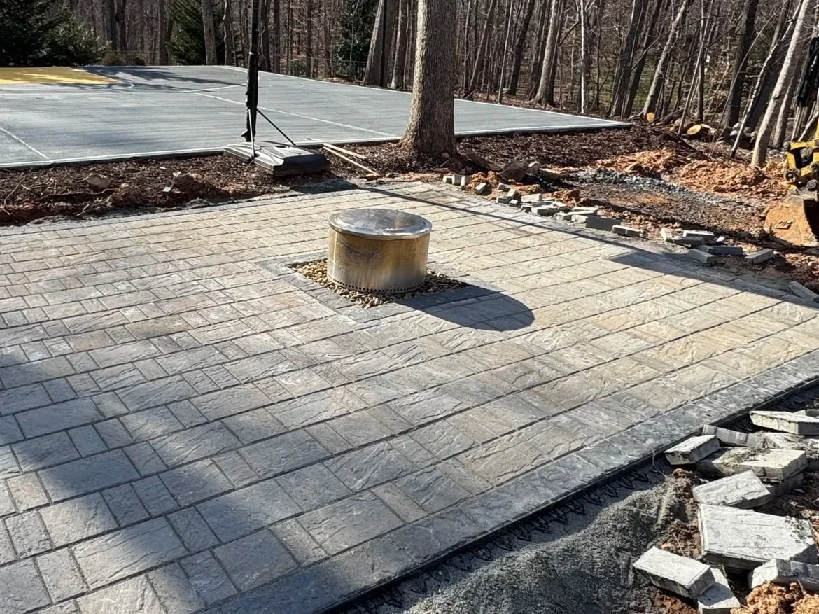 A partially completed stone patio with a silver vent pipe in the center, a tree, and a paved basketball court in the background, surrounded by trees and construction materials.