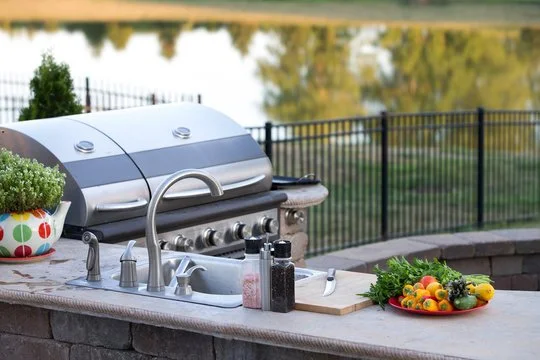 Outdoor kitchen counter with a gas grill, potted plant, salt and pepper shakers, a cutting board, and a bowl of colorful vegetables and greens, overlooking a fenced yard and water body.