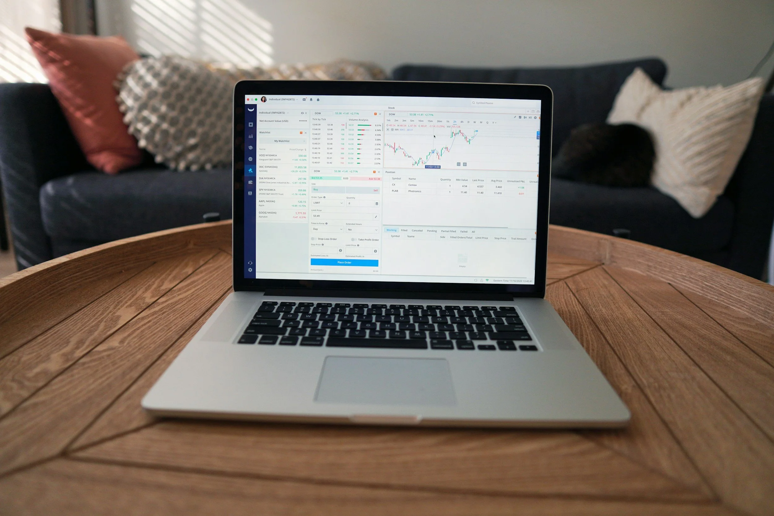 A laptop displaying stock market charts and trading information on a round wooden table in a cozy living room with cushions on a sofa in the background.