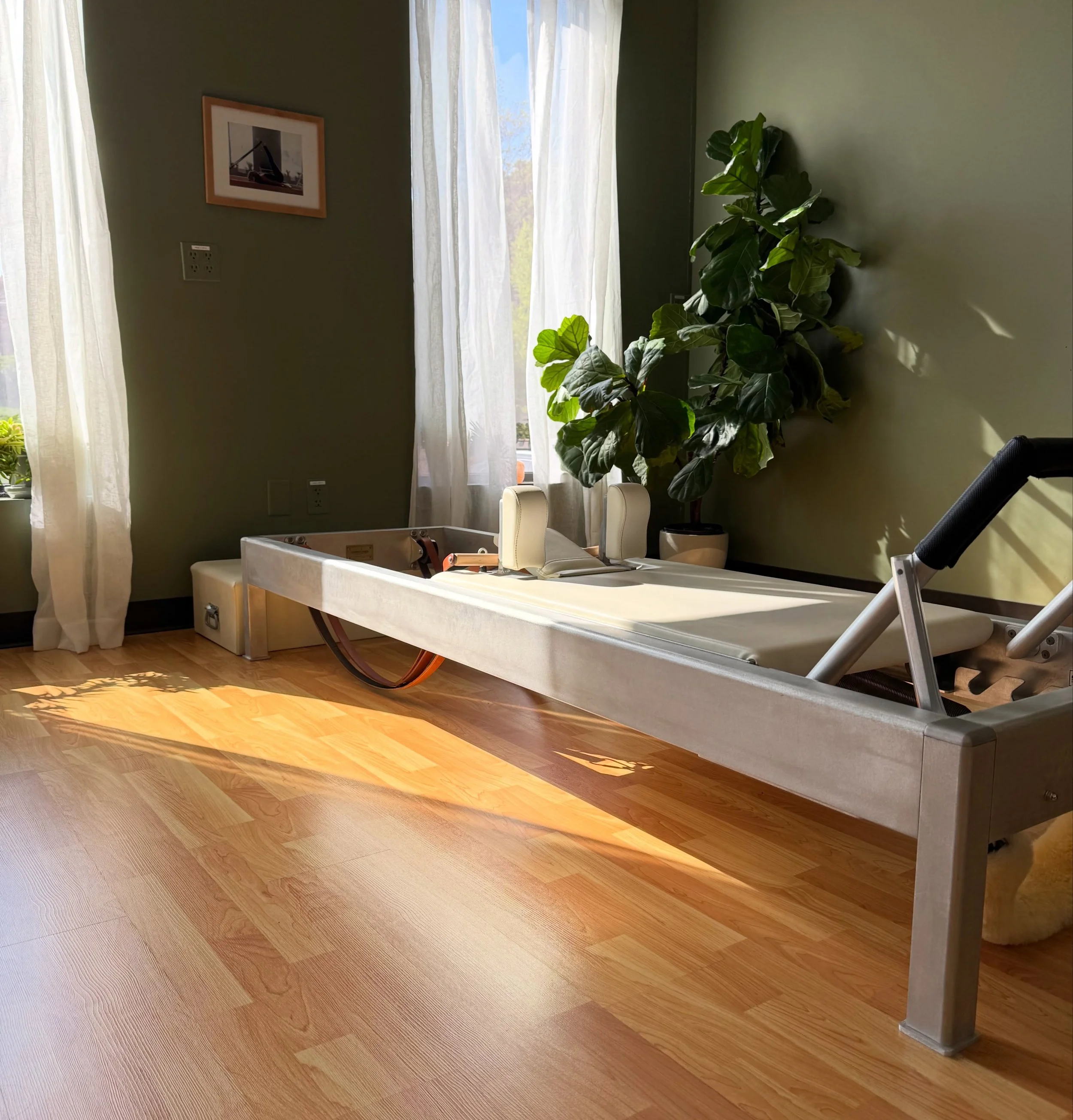 Sunlit room with wooden flooring, a large green houseplant in a white pot, white curtains covering a window, and a wall with a framed picture of a steel structure. There is a foosball table with white and black seats, and part of a black office chair is visible.