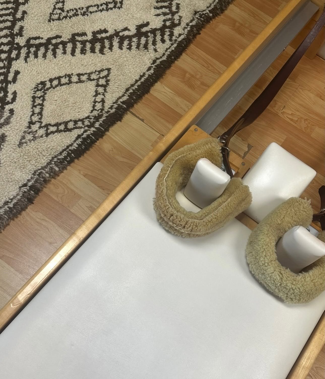 Close-up of pilates reformer with sheepskin covered ankle bands. A patterned rug on a wooden floor is partially visible.