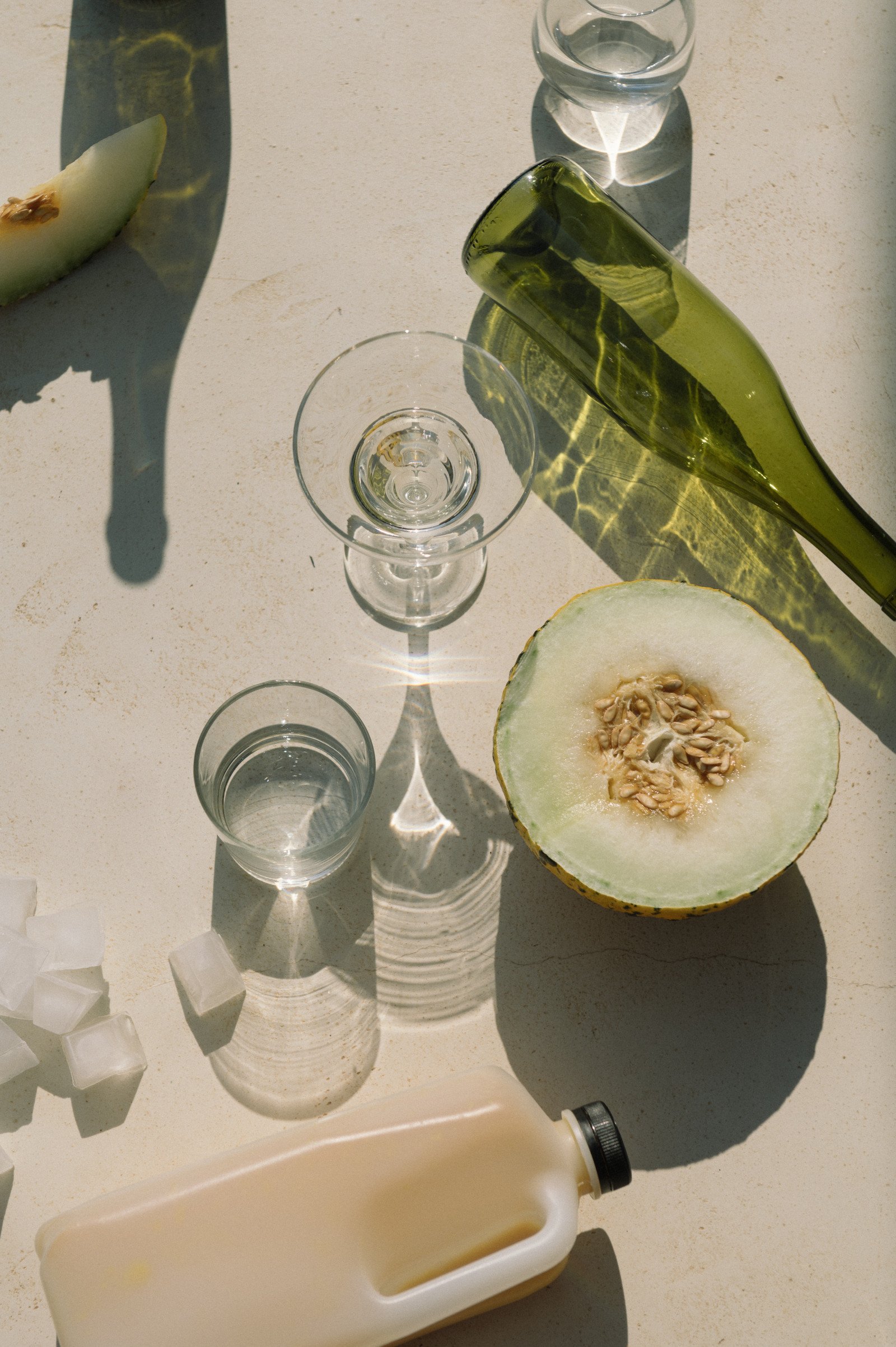 A variety of objects on a light-colored table, including a halved cantaloupe with seeds, a glass of water, a small empty glass, a container of light yellow liquid, ice cubes, a green glass bottle, and a bottle of white liquid.