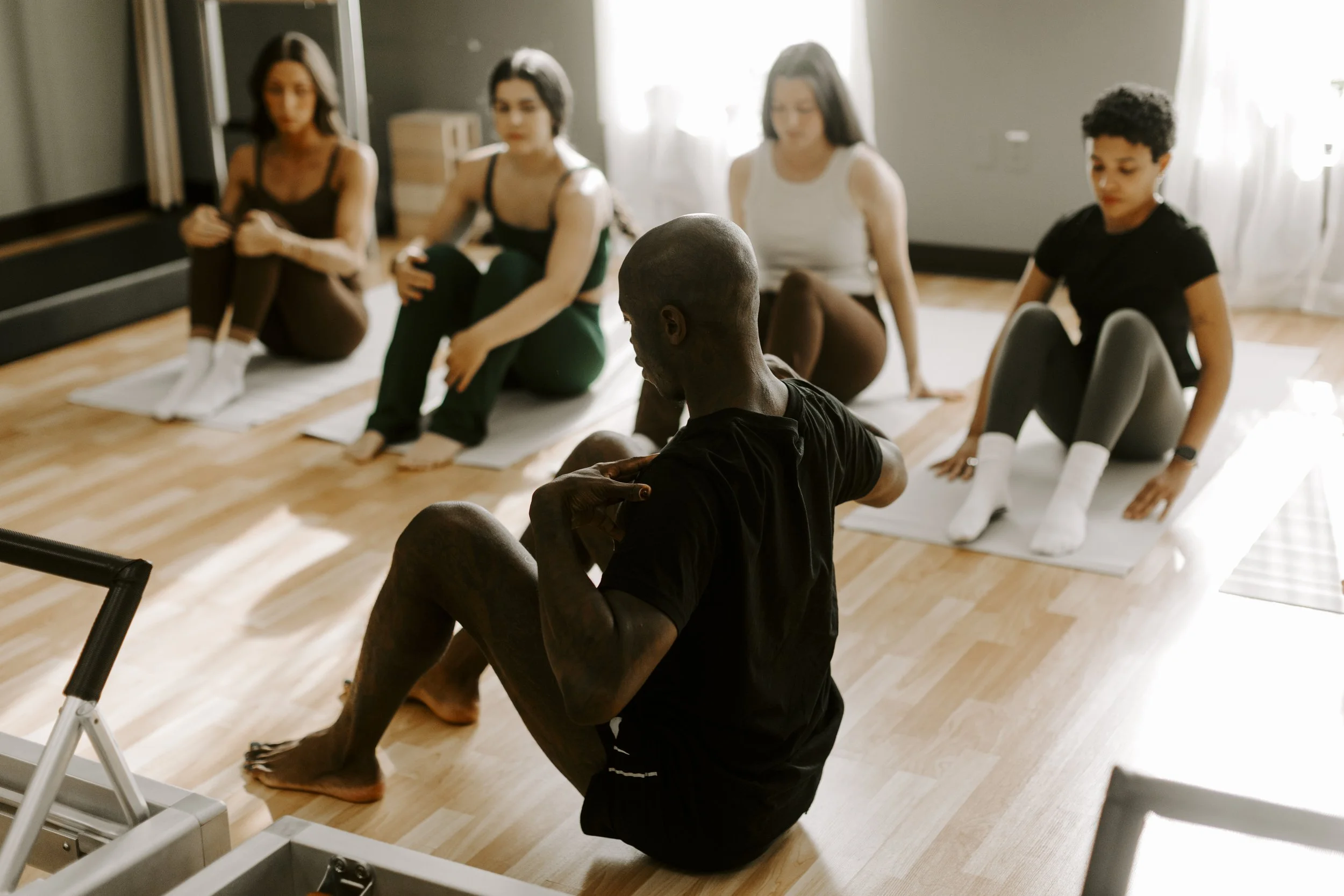 A group of five people, four women and one man, are sitting on yoga mats in a room, appearing to be in a meditation or mindfulness practice. The man in the foreground is seated cross-legged with his eyes closed, while the women in the background are also seated on mats, with seated postures and closed eyes.