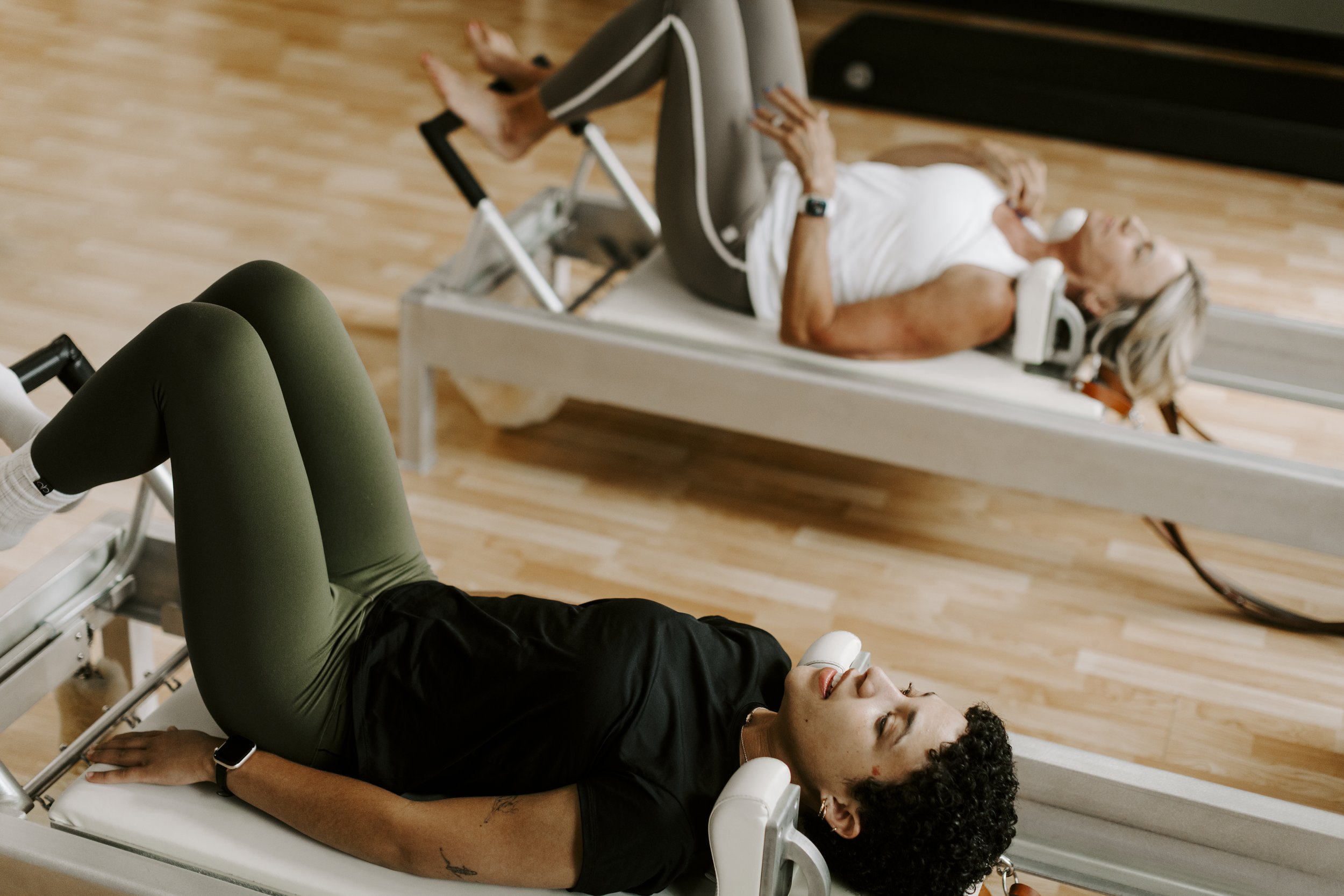 A woman lying on a pilates reformer with her eyes closed, with another woman in comfortable workout attire laying on a reformer nearby in an exercise studio with wooden floors.