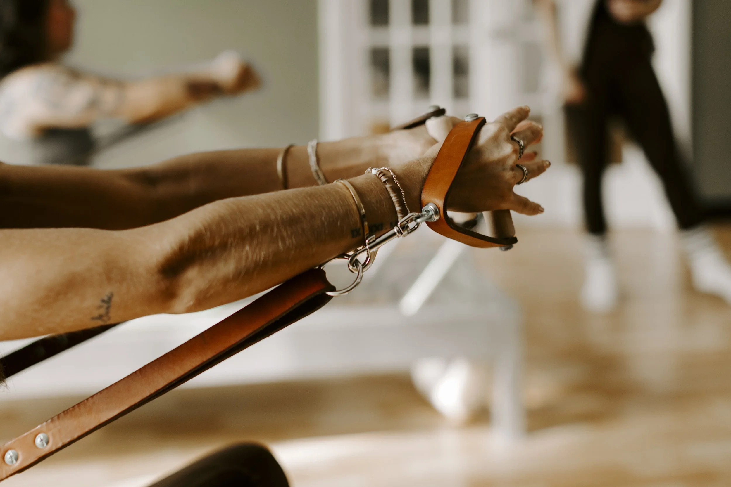 Close-up of a person's arms and hands with bracelets, rings, and tattoos, holding leather straps connected to a metal harness of a pilates reformer, with people in the background.