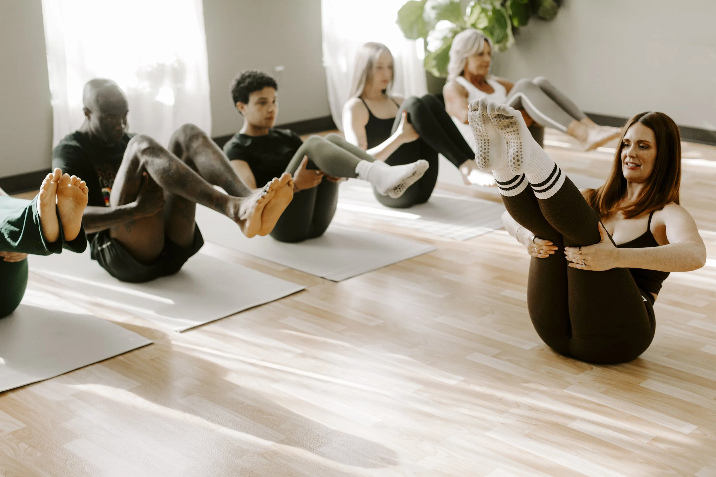 A group of people participating in a mat pilates class, lying on yoga mats while a instructor helps a woman with her pose.