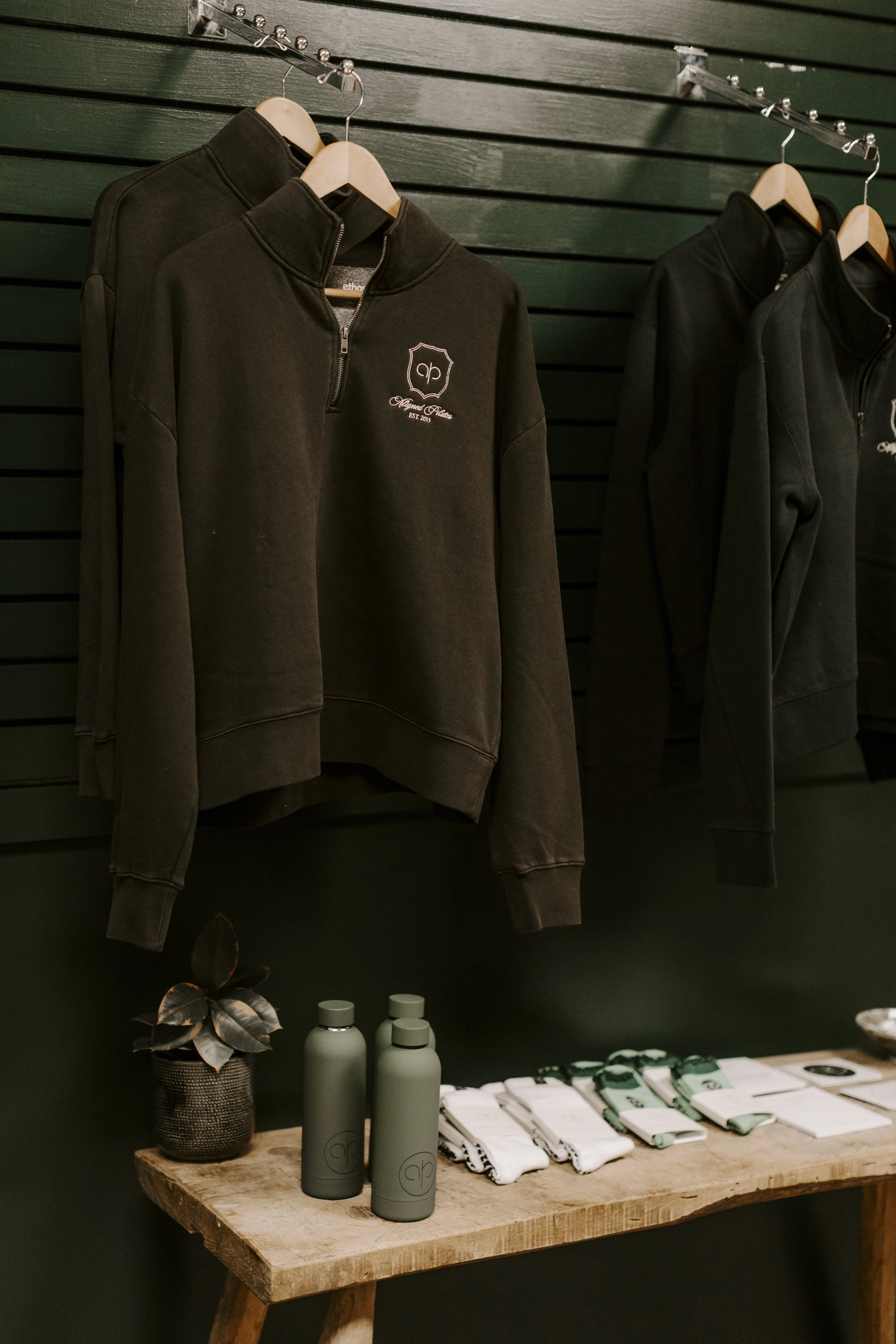 Display of black sweatshirts with a logo on hangers, bottles and merchandise on a wooden table, plant in a basket, against a dark green slatted wall.
