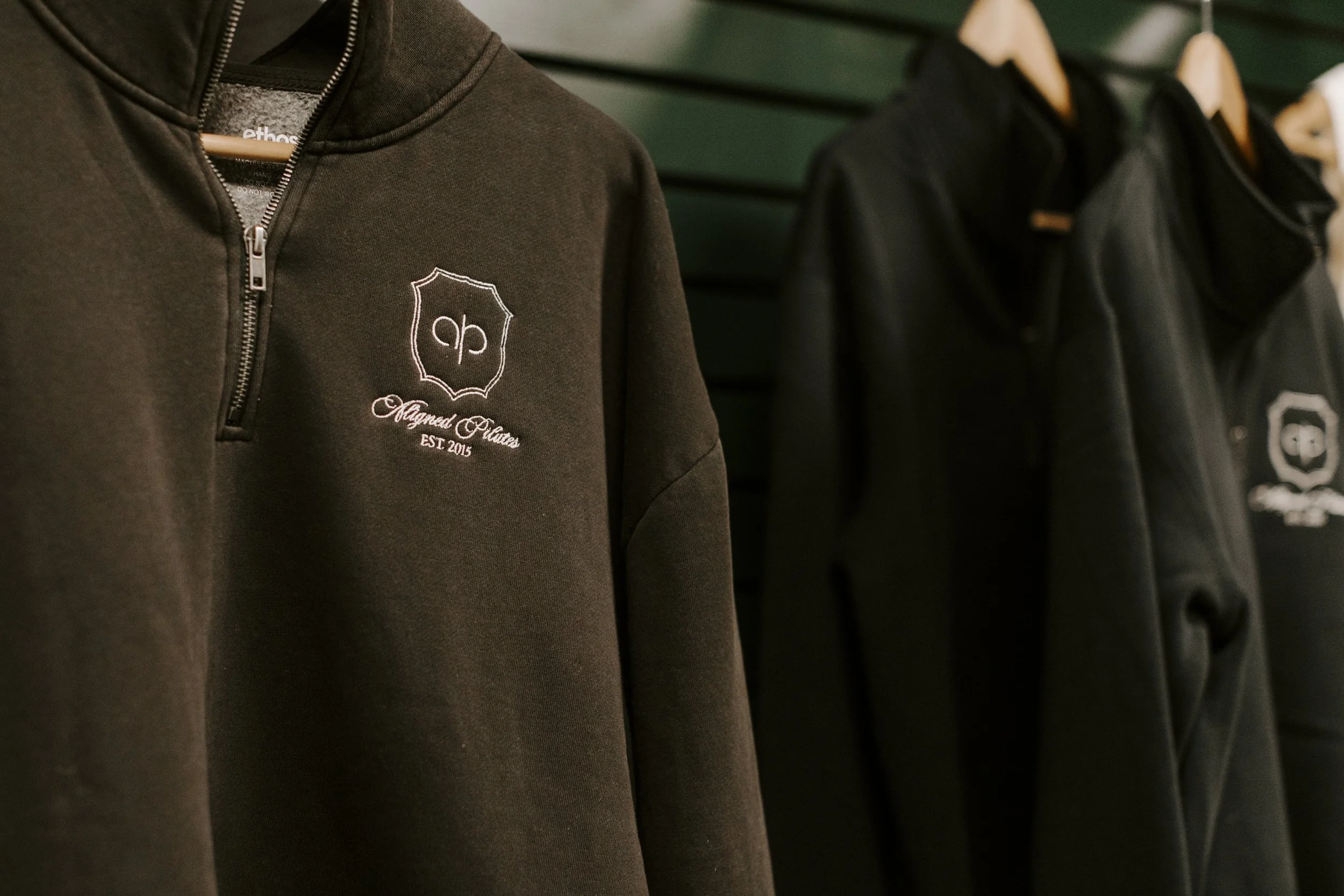 Close-up of a black hoodie with a logo and text on the chest, hanging on a hanger, with other black jackets hanging on hooks in the background.
