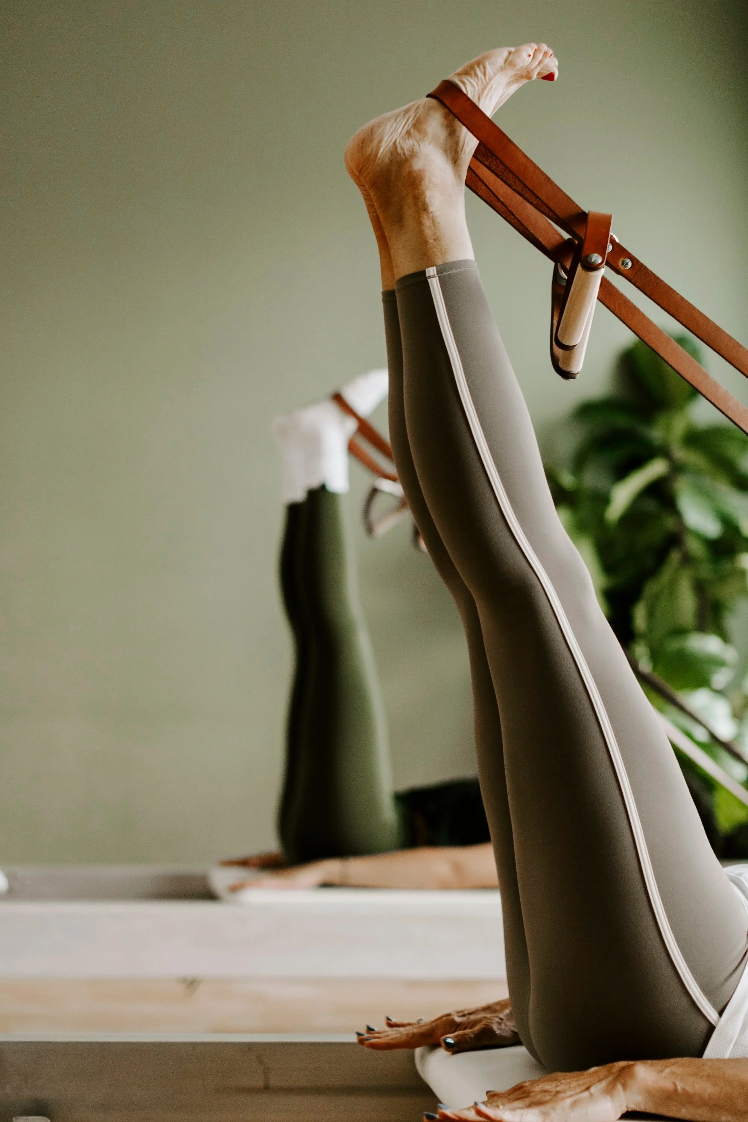 Close-up of a person's legs and feet in pilates move, with hands on the reformer machine in a room with a green wall and a houseplant in the background.