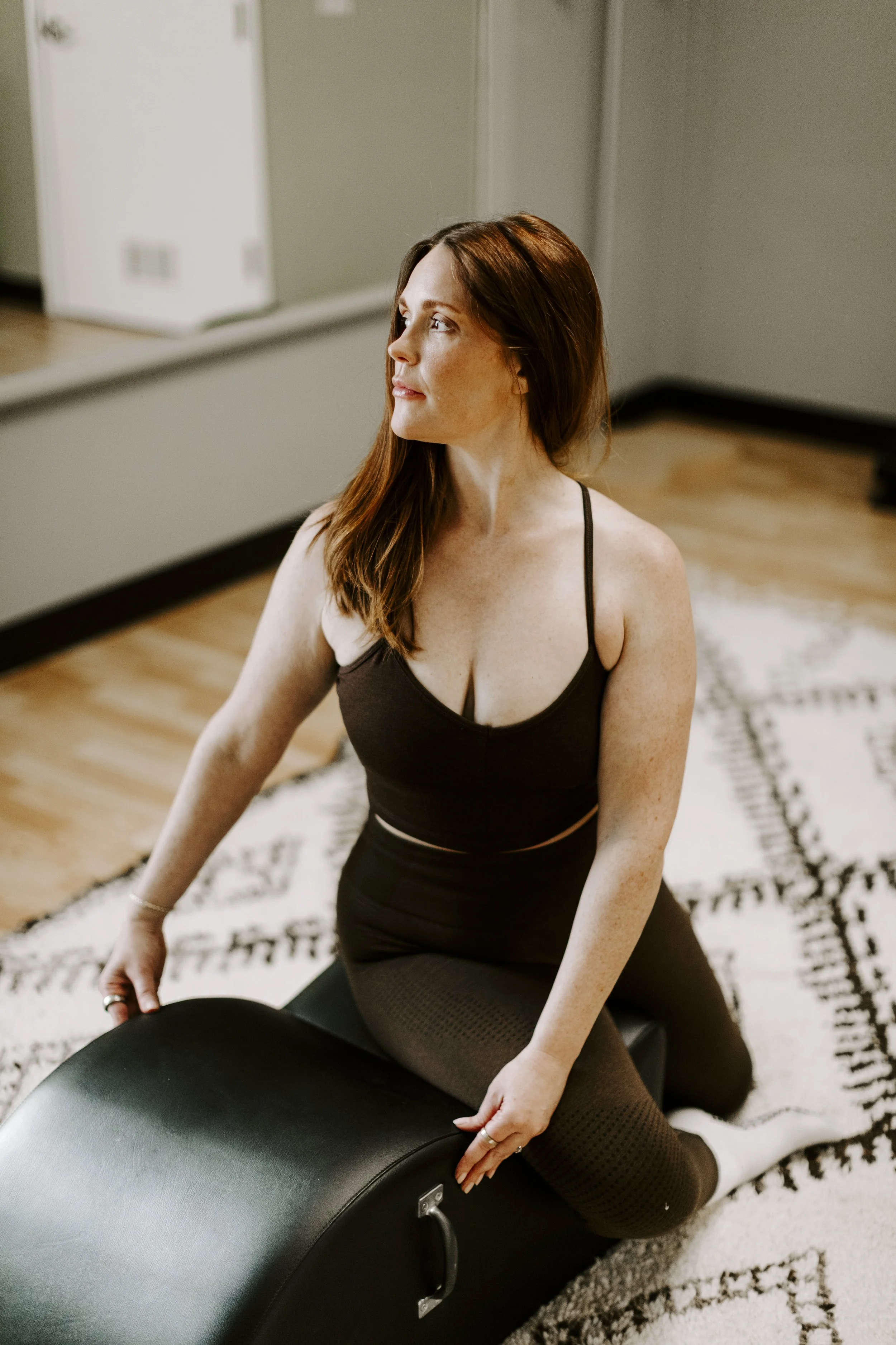 A woman with long red hair practicing pilates in a room with a mirror, beige walls, and hardwood floors. Woman is seated on a textured rug.