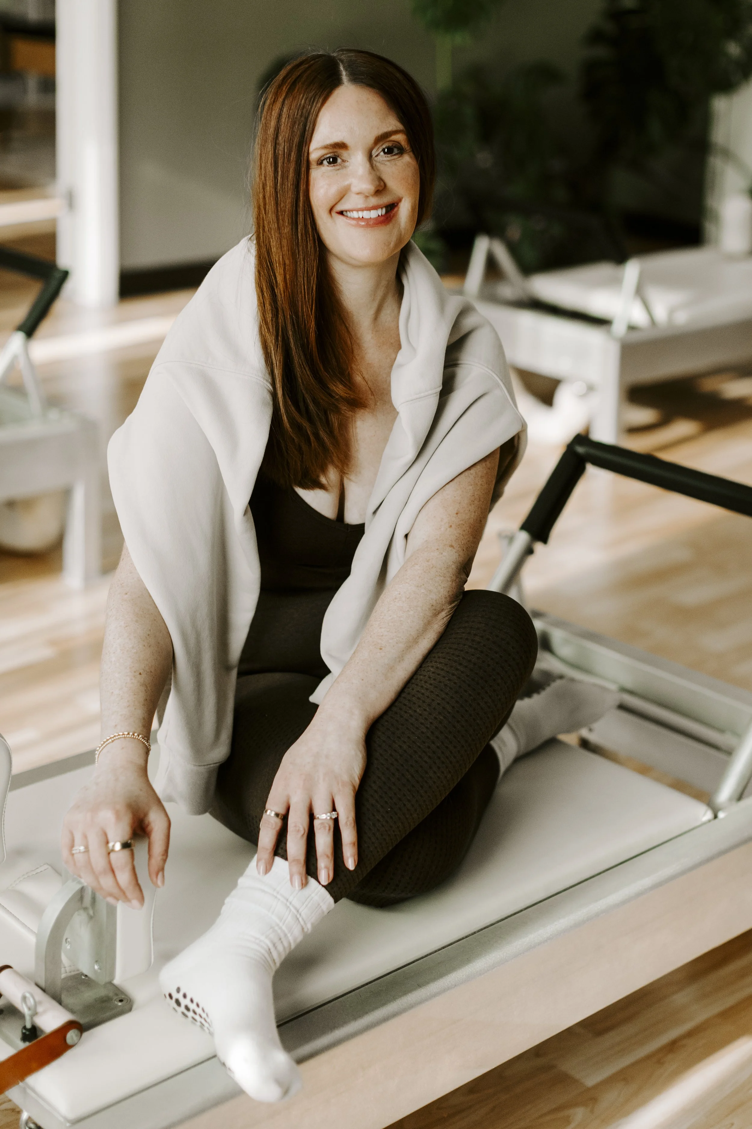 A woman with red hair sitting on a pilates reformer posing for a formal portrait with plants in the background.