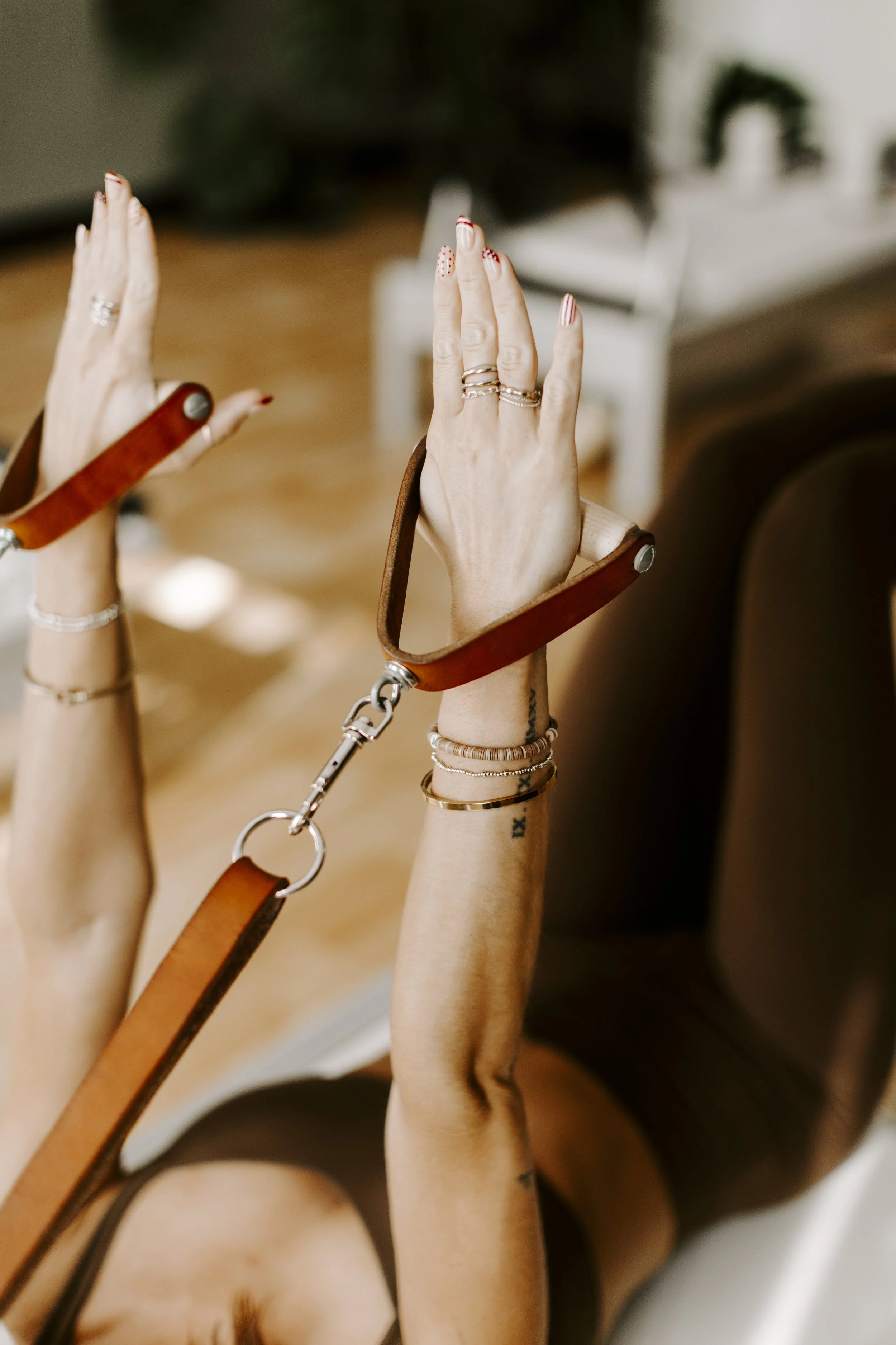 A person with their hands raised, wearing multiple rings, and bracelets holding reformer bands in a stretch.