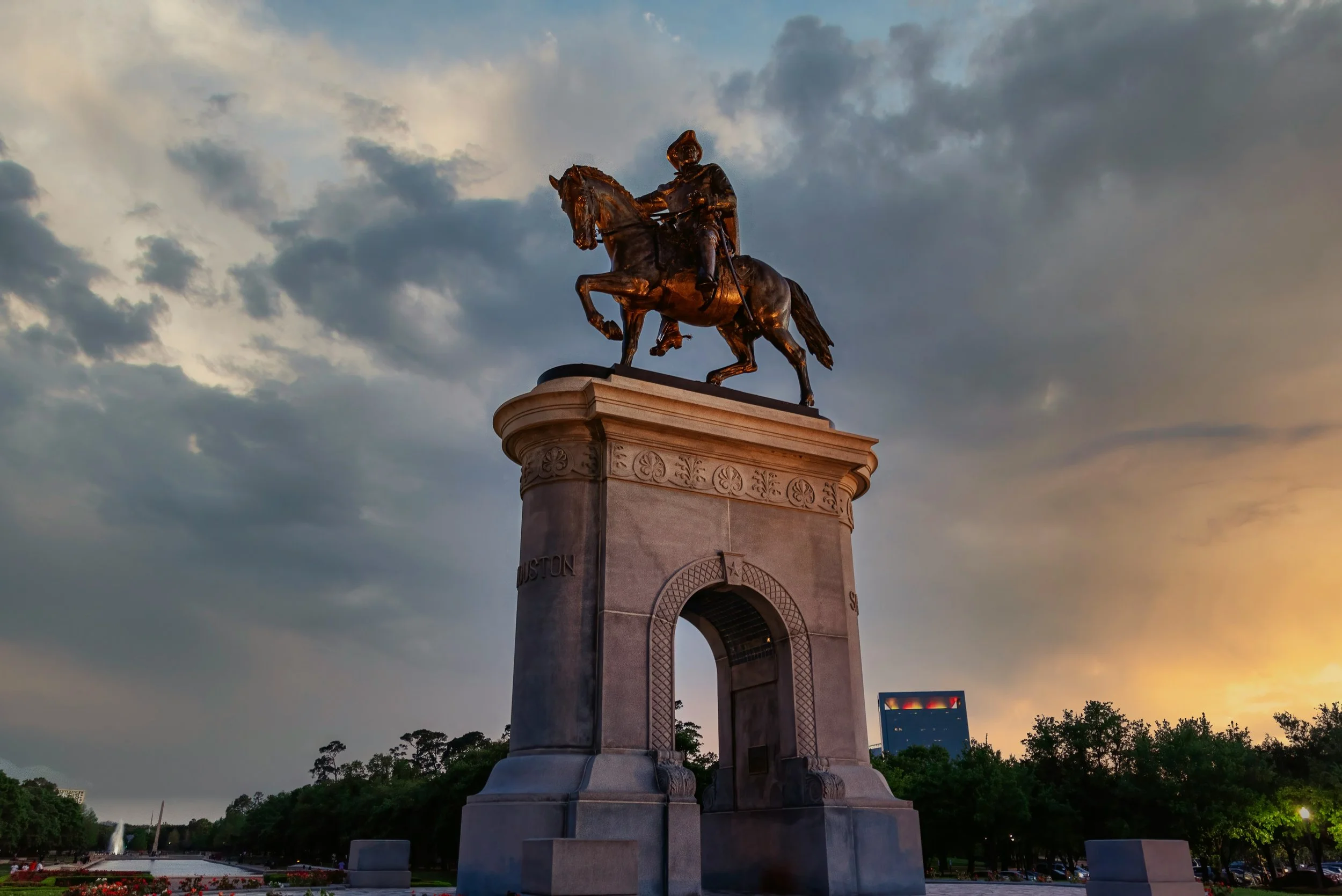 Bronze statue of a man on horseback mounted on a tall stone pedestal, set against a cloudy sky during sunset.
