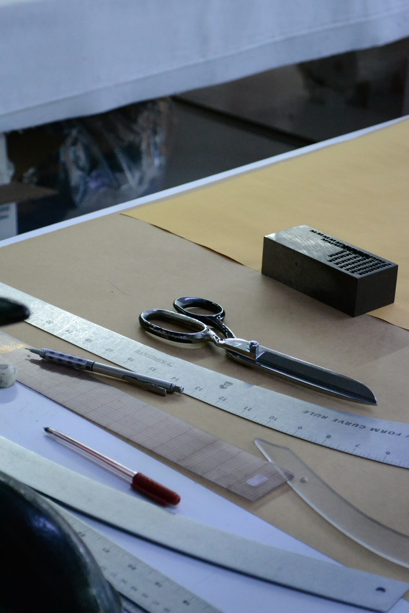 A work surface with scissors, a ruler, a pen, and a metal stamp block, with a sheet of paper and yellow sheet, along with other miscellaneous items.