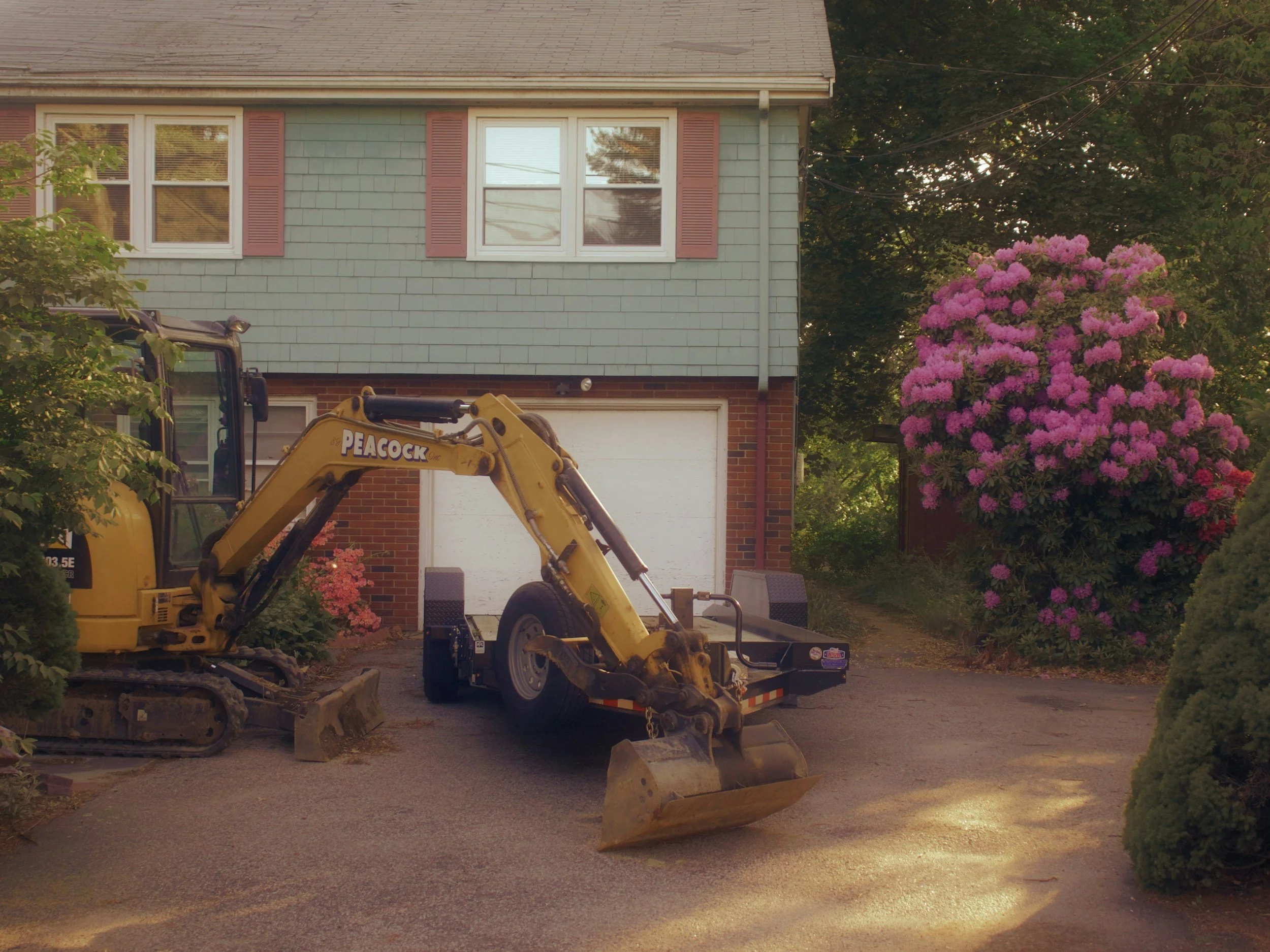A yellow construction excavator parked in front of a house with light green siding and red shutters. There is a large pink flowering bush and greenery around the house, with overhead power lines in the background.