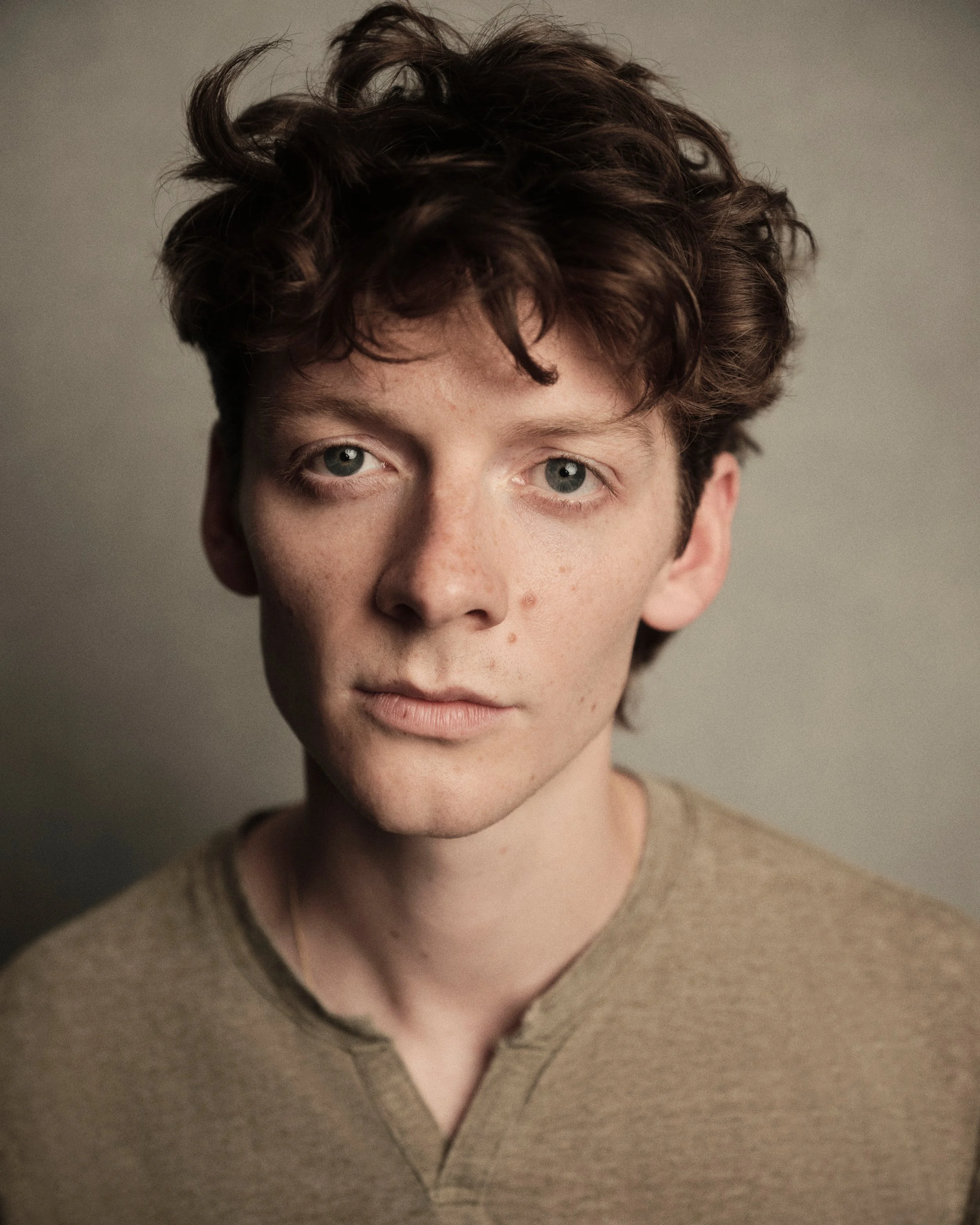 Close-up photo of a young man with curly brown hair, blue eyes, and fair skin, wearing a beige V-neck sweater.