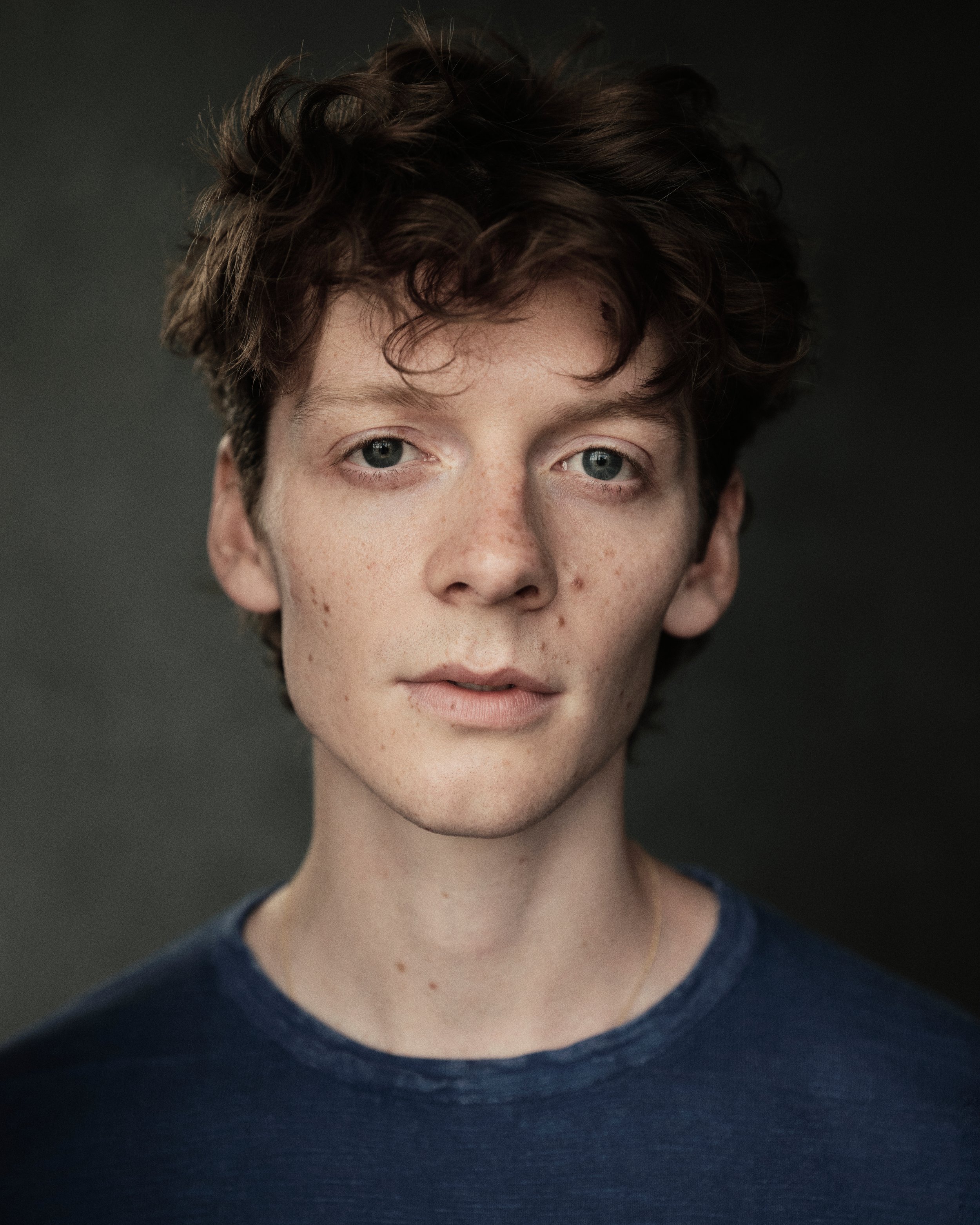 Close-up of a person's face with short, curly brown hair, fair skin, and wearing a dark blue shirt.
