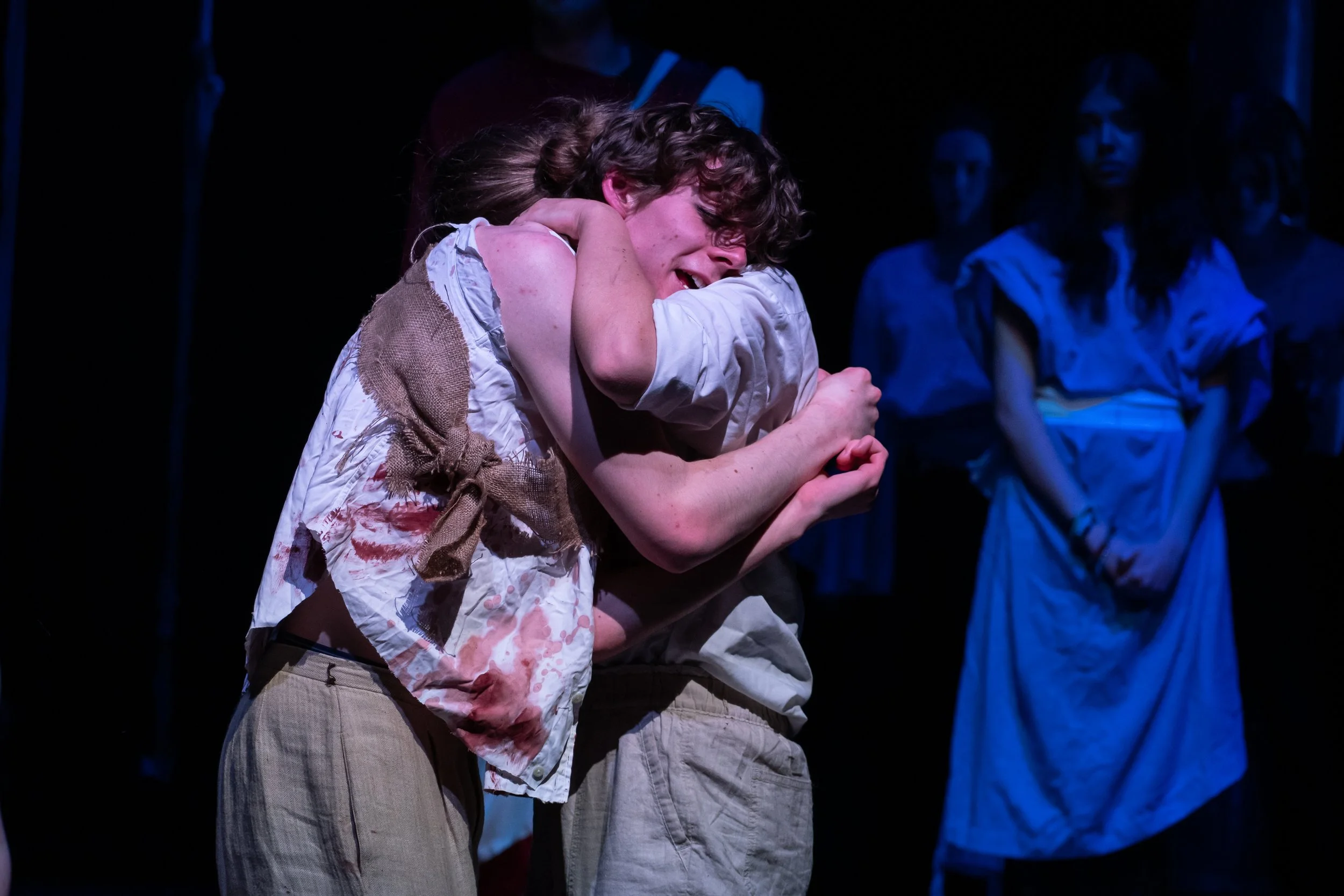 Actors on stage performing a scene involving an emotional embrace, with some actors in the background observing.