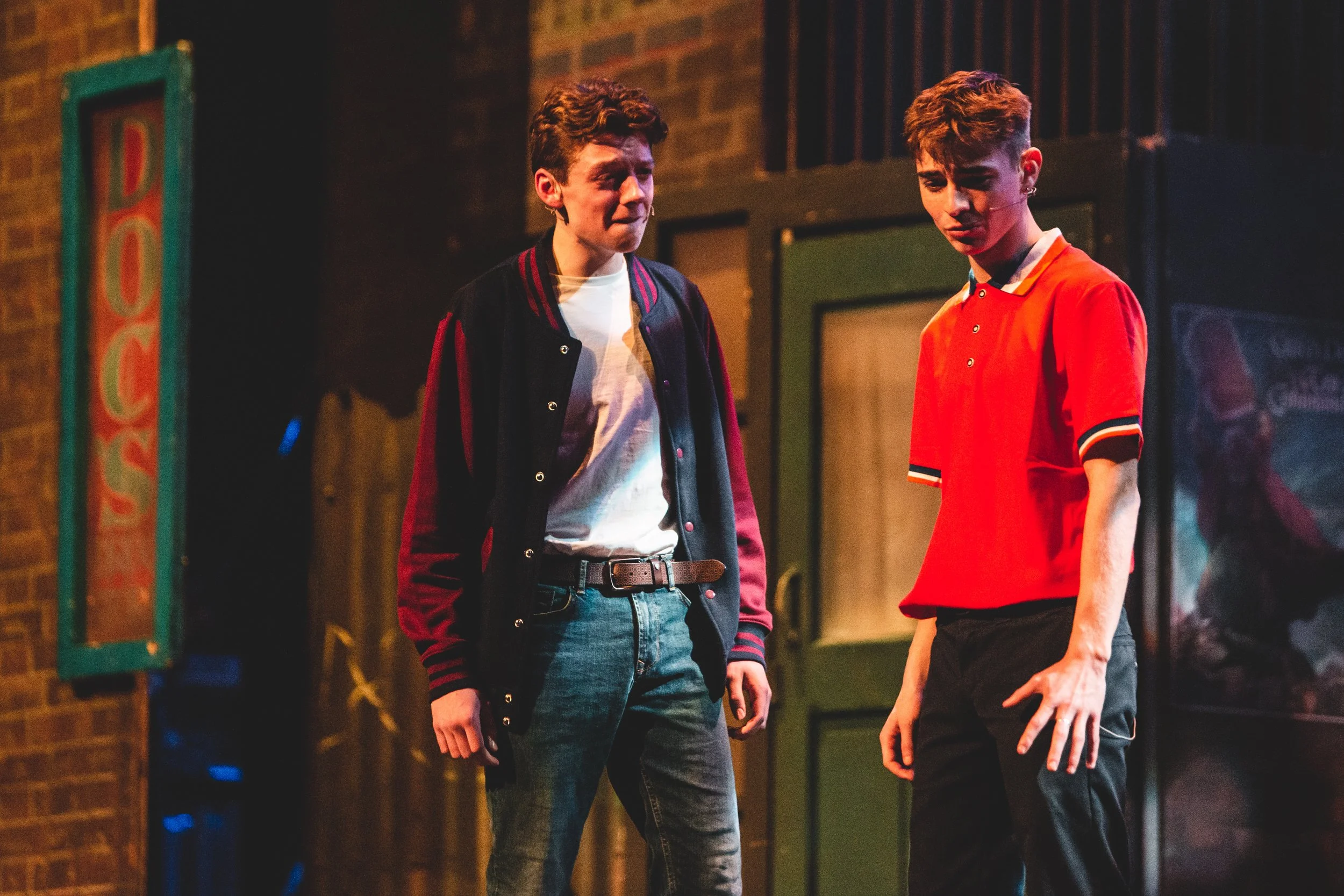 Two young men standing in a dimly lit indoor setting, possibly on stage, engaging in a conversation or performance, with one wearing a black and red jacket and the other in a red polo shirt. The background includes brick walls and framed artwork.
