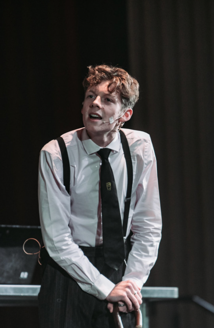 A young man with curly brown hair and light skin, wearing a white dress shirt, black tie, suspenders, and dark pants, is standing on a stage speaking into a headset microphone, with a dark background and some equipment behind him.