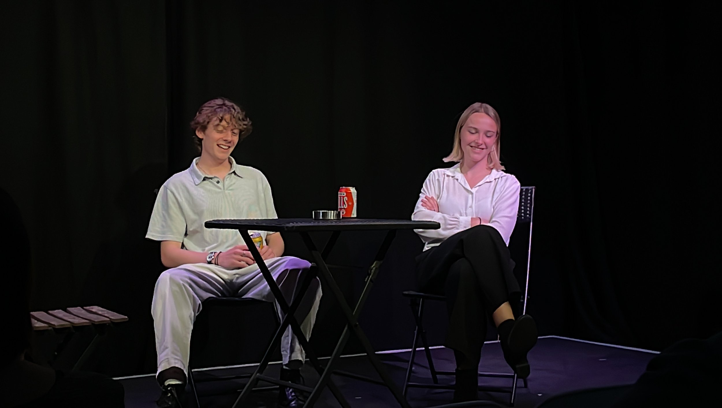 Two people sitting at a table on a stage. One person is a young man with curly hair, wearing a light-colored shirt and pants, holding a can of soda. The other person is a young woman with straight blonde hair, wearing a white blouse and black pants, 