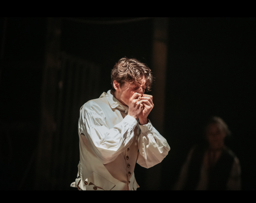 A man with curly hair wearing a white shirt, appears to be emotionally distressed, covering his face with both hands, in a dimly lit setting.