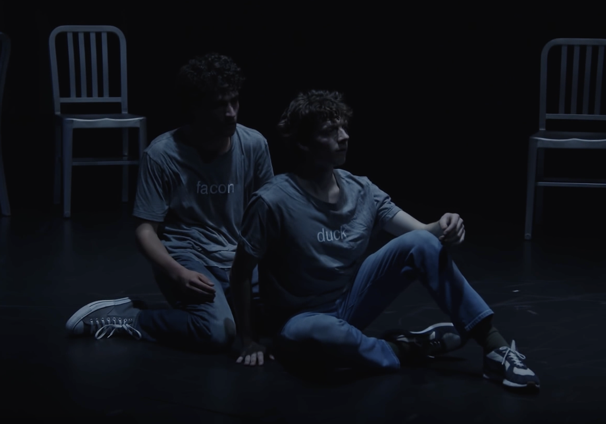 Two young men are sitting on the floor in a dark room with a black background, surrounded by empty chairs. They are both wearing casual clothing and appear to be engaged in a serious or emotional moment.
