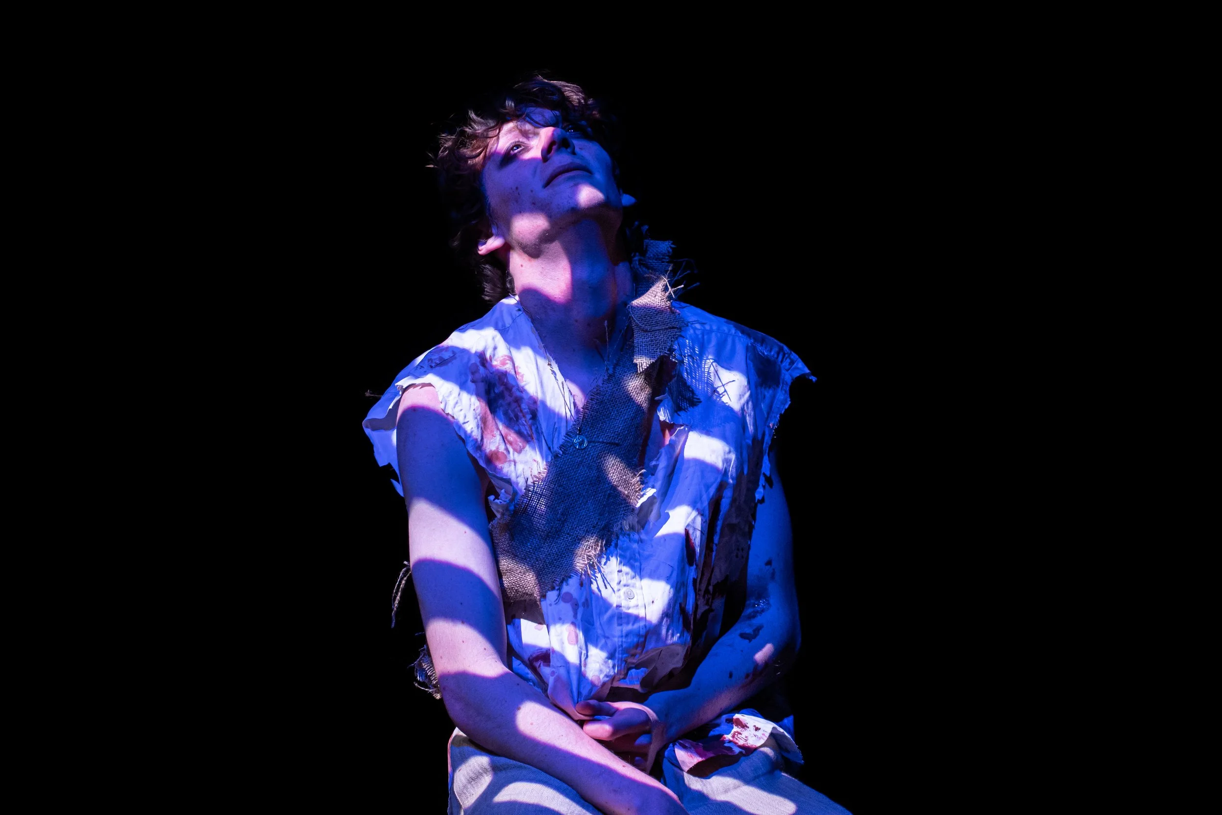 A person with curly hair looking upwards, illuminated by blue light, wearing a torn, patterned shirt, against a black background.