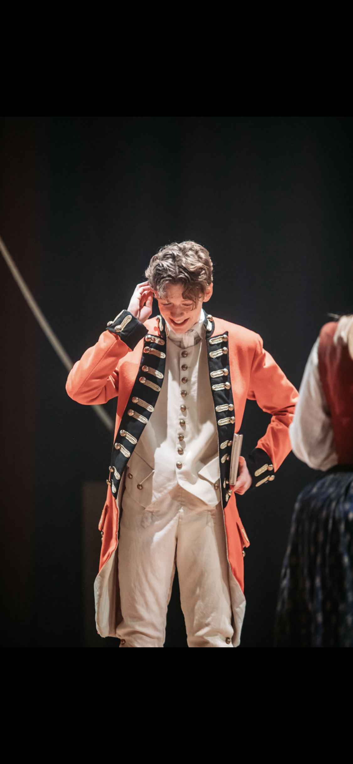 A young man in historical military costume, with a red coat and white trousers, smiling and scratching his head, standing on stage.