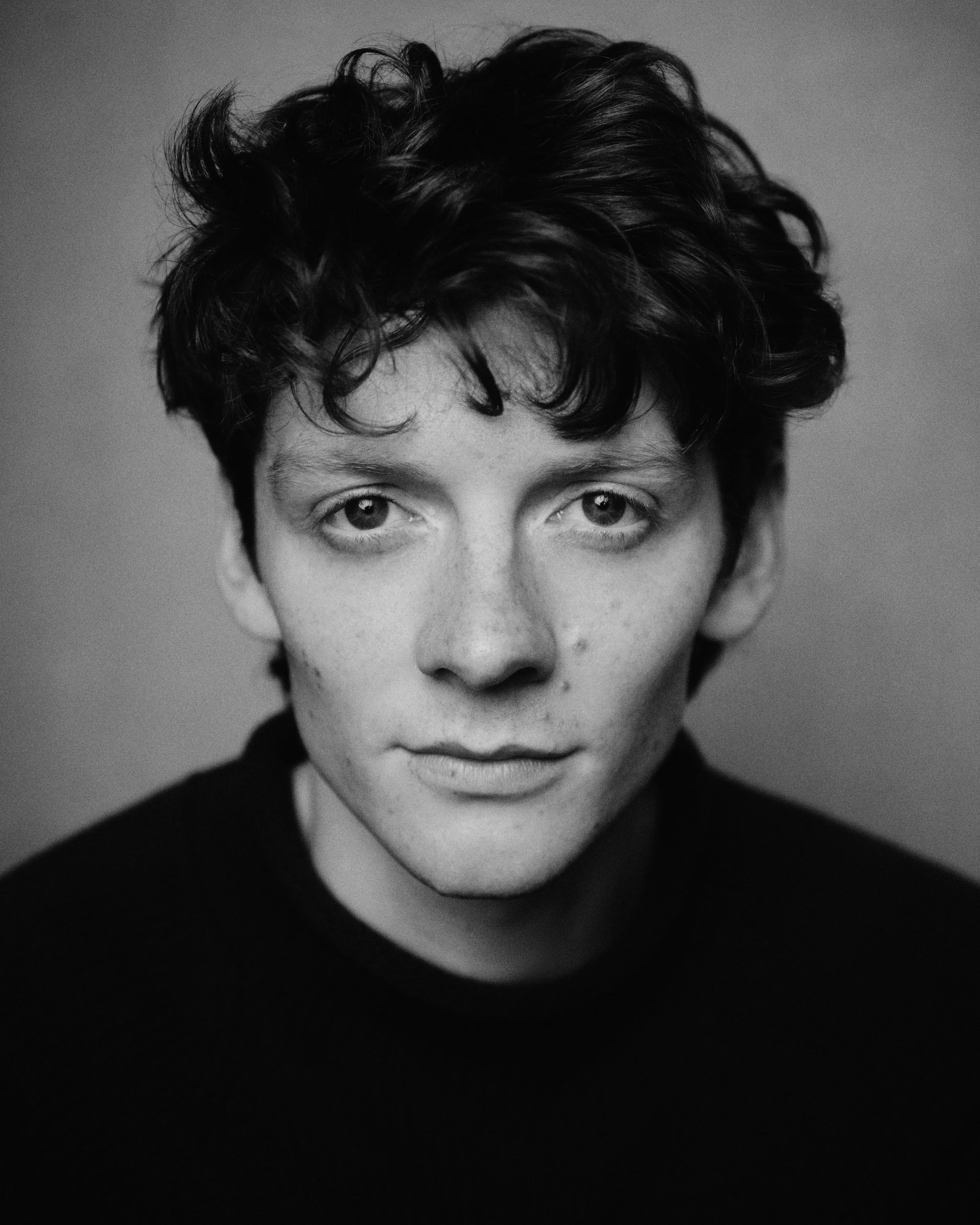 Black and white close-up portrait of a man with short, curly hair, staring directly at the camera. He is wearing a dark top and has a neutral expression.