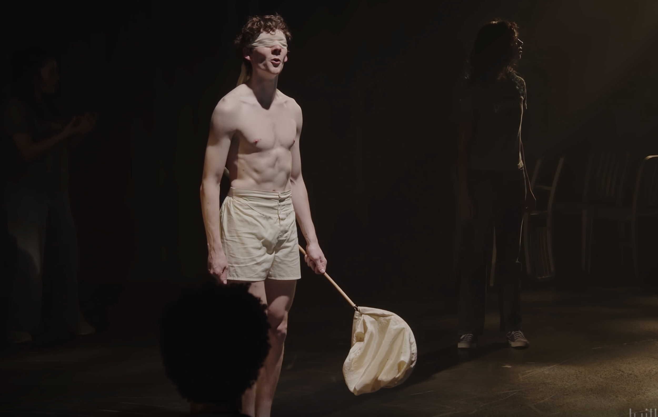 A shirtless man with a blindfold over his eyes, wearing beige shorts, stands on a stage holding a stick. There are other people in the background, seated and standing in the dark.