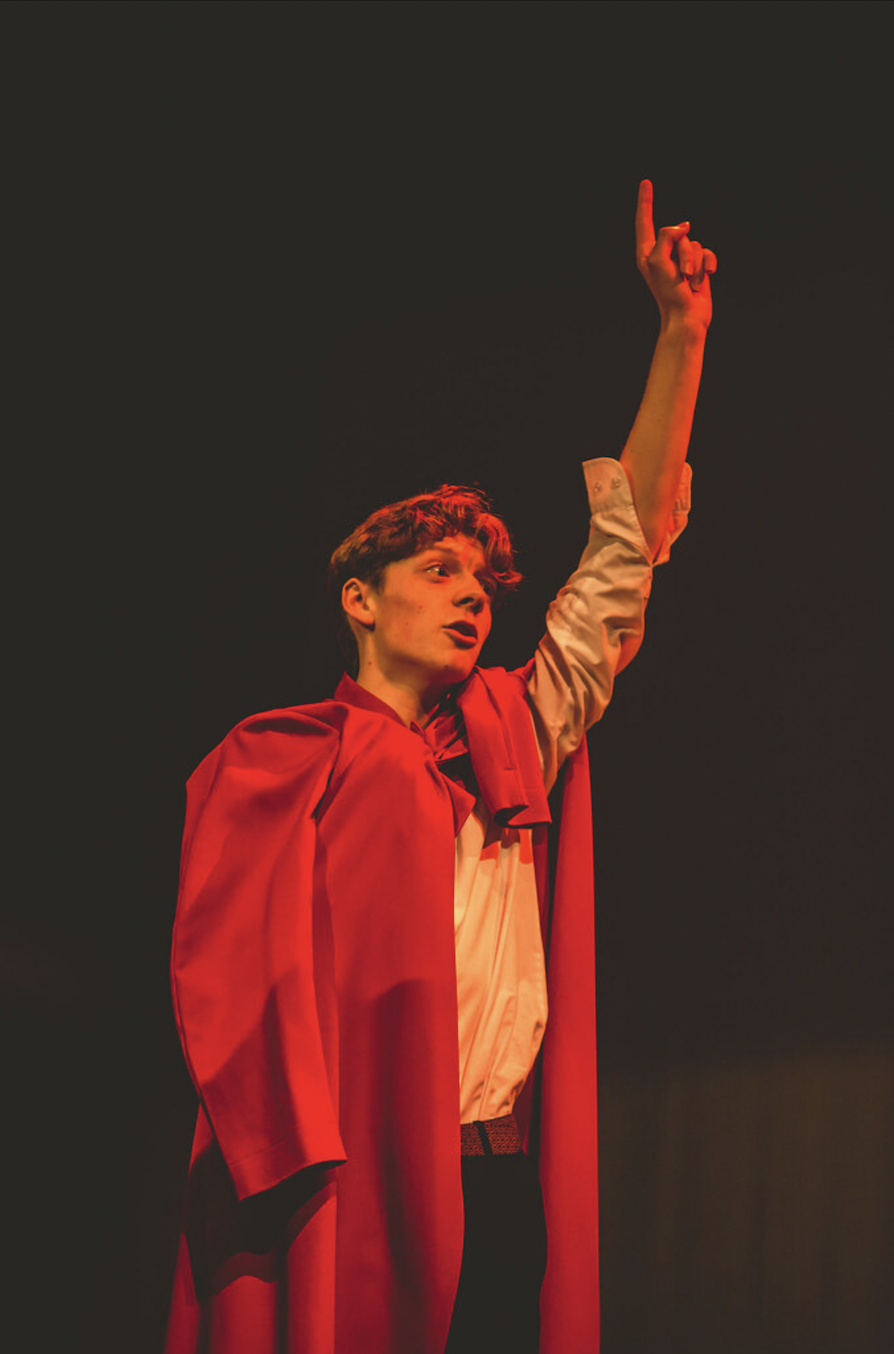 A young man wearing a red cape and white shirt, gesturing upward with his right hand, on a dark stage with dramatic lighting.