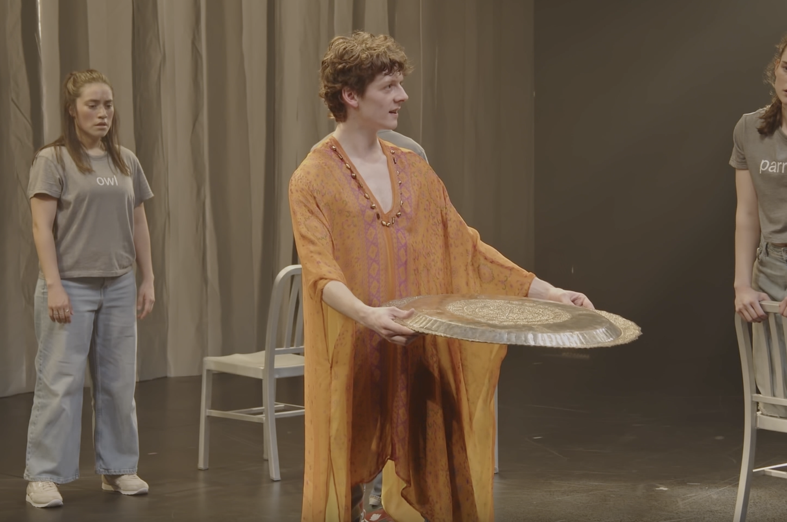 A man in an orange kaftan holding a large, round, shiny object in a room with beige curtains and two young women standing nearby, one on each side.