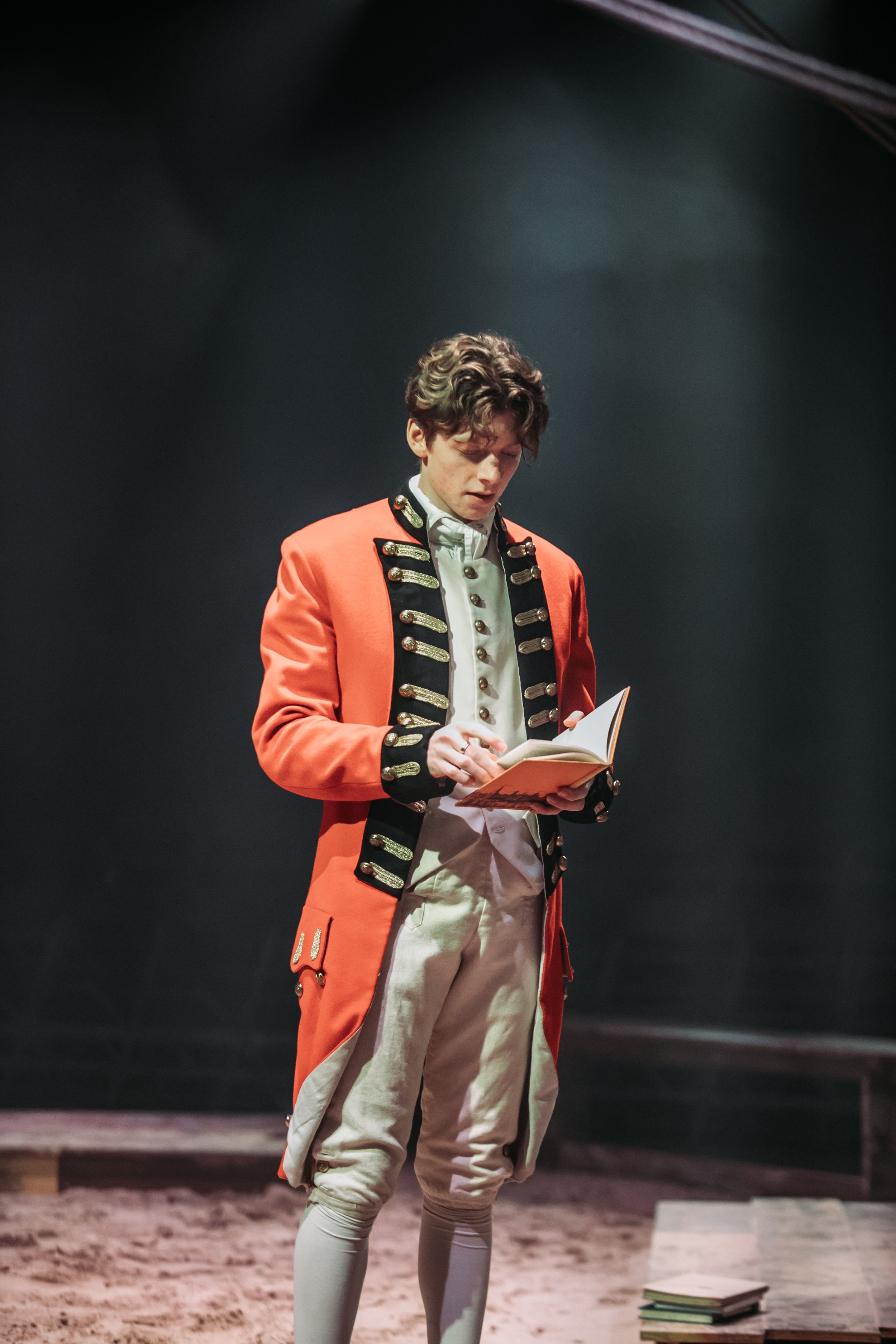 Young man dressed in historical military uniform reading a book on a stage. There are several stacks of books on the stage floor beside him.