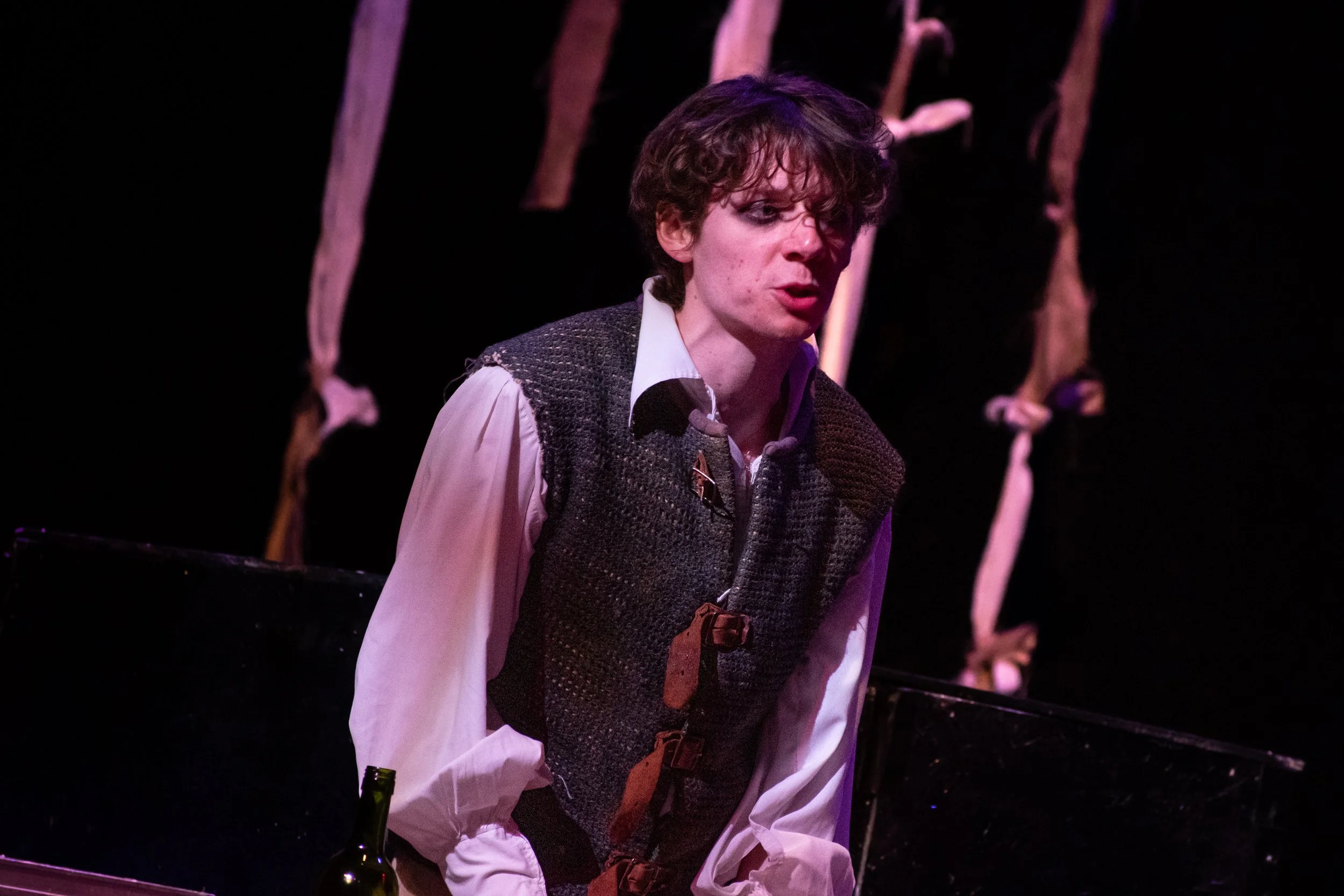 A person with curly hair wearing a white shirt and a vest, looking contemplative with dark makeup, on stage with what appears to be a theatrical background.
