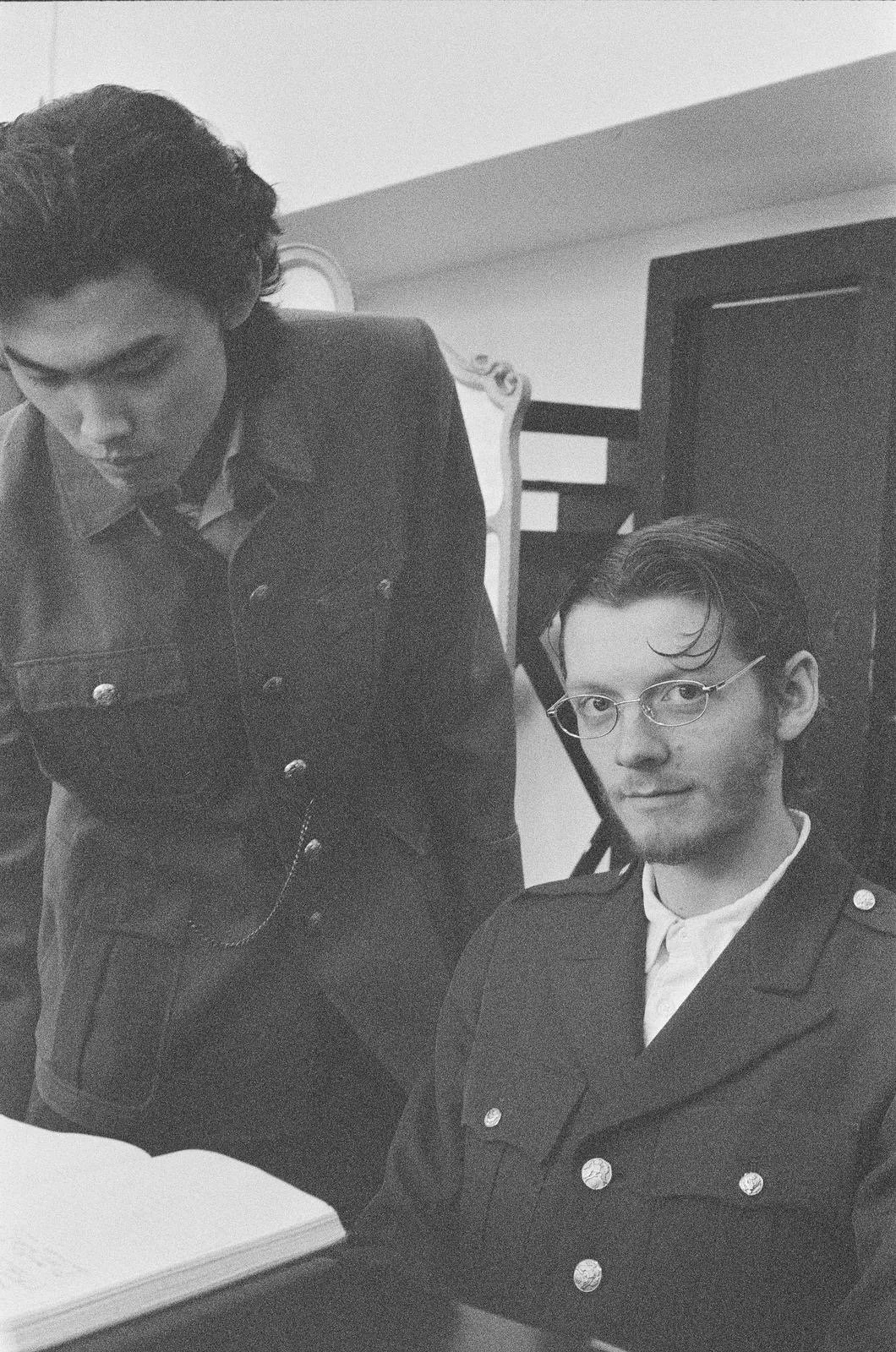 Two men, one standing and one seated, in black jackets and glasses, in a room with a staircase and a closed door in the background