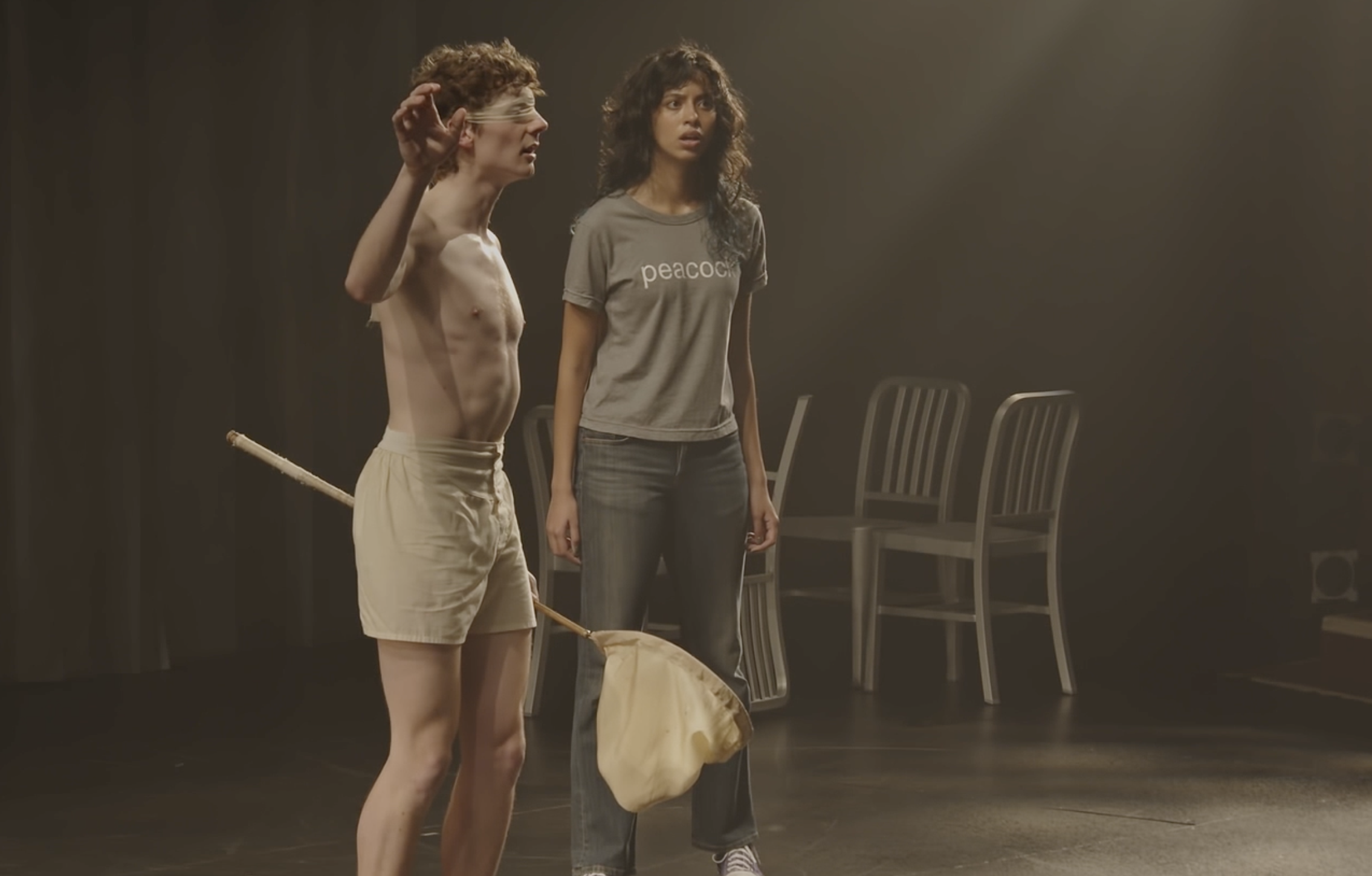 A shirtless man with a blindfold tied around his eyes and beige shorts holding a stick stands next to a woman with curly dark hair, wearing a gray "peacock" t-shirt and jeans, in a room with chairs in the background.