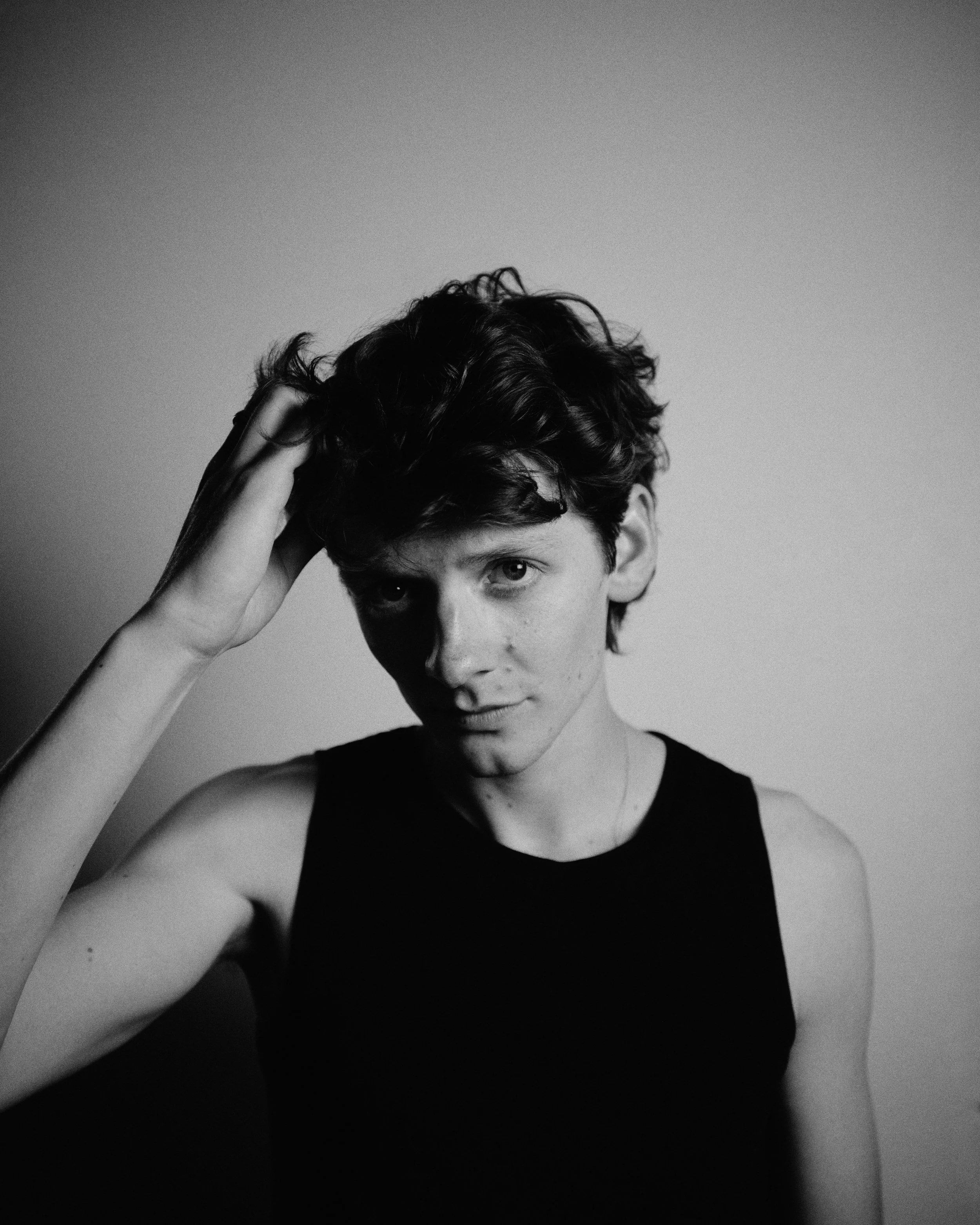 Black and white photo of a young man with short, curly hair, wearing a sleeveless top. He is touching his head with his right hand and looking directly at the camera with a slight smile.