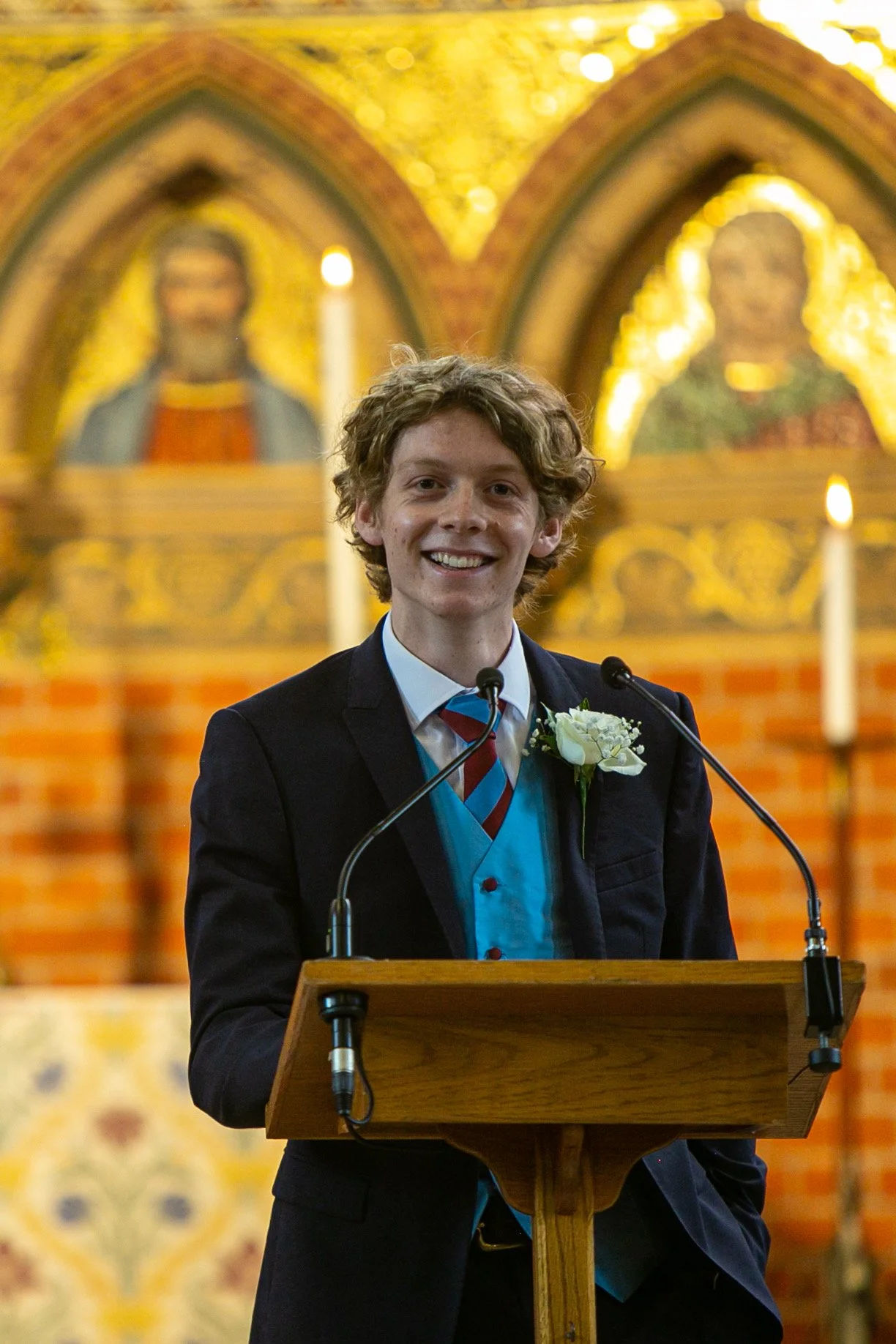A young man in a formal suit and tie, standing at a wooden podium with a microphone, smiling in a church, with religious paintings and lit candles in the background.