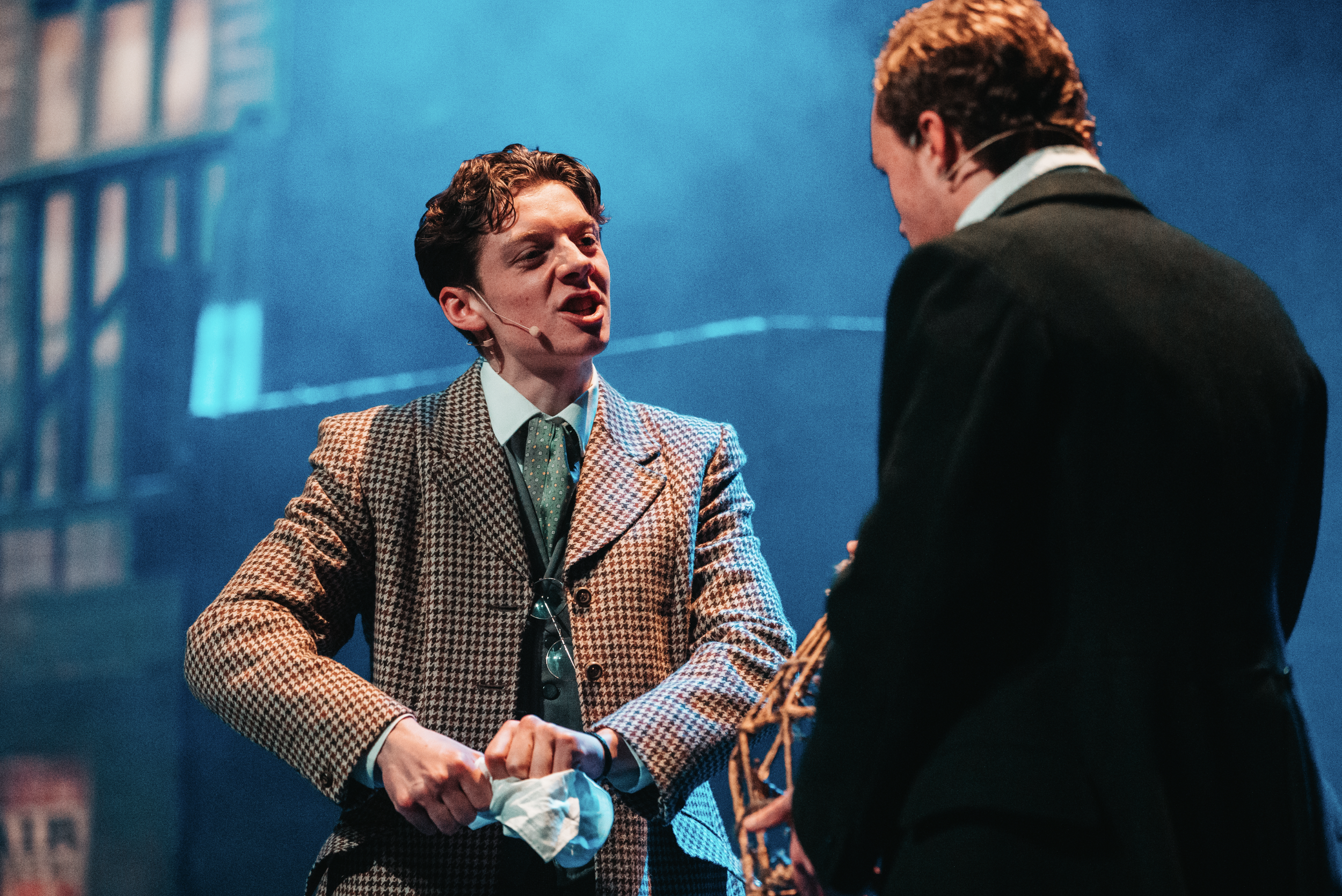 Two men shaking hands on stage during a presentation or speech, with one wearing a checkered blazer and the other in a dark suit, against a blue background.