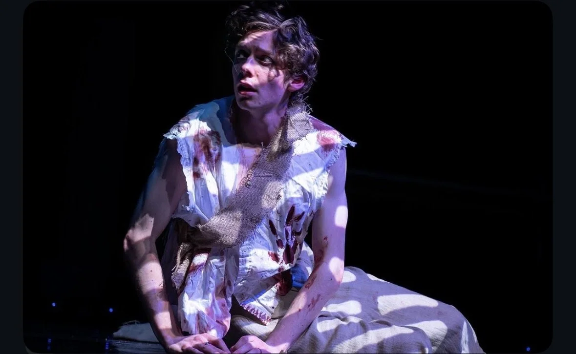 Person with messy hair and torn, blood-stained clothing sitting on stage in a dark environment, lit by dramatic lighting.