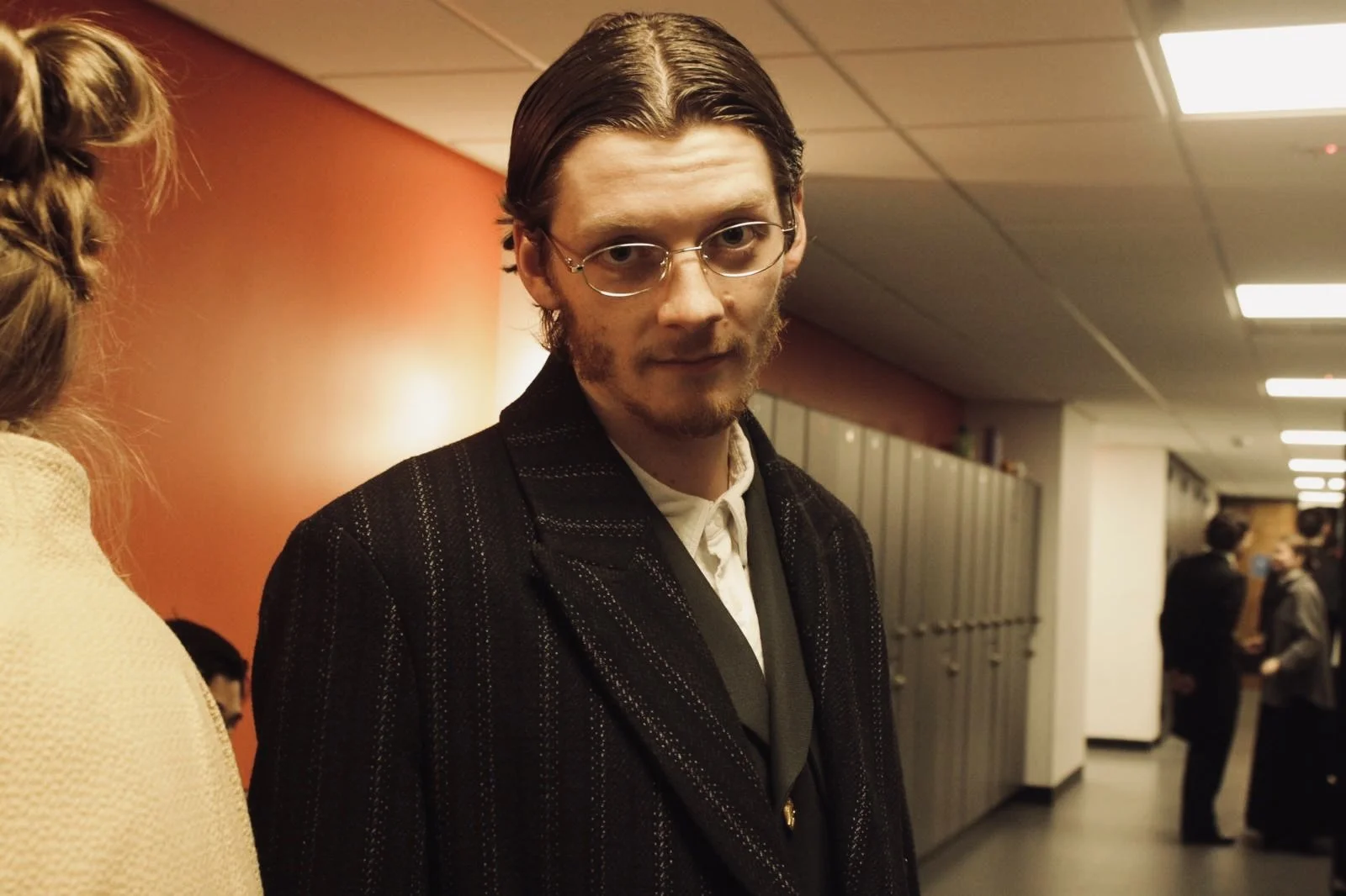 A young man with glasses and facial hair looking at the camera in a hallway with lockers. There are other people in the background, some standing and talking.