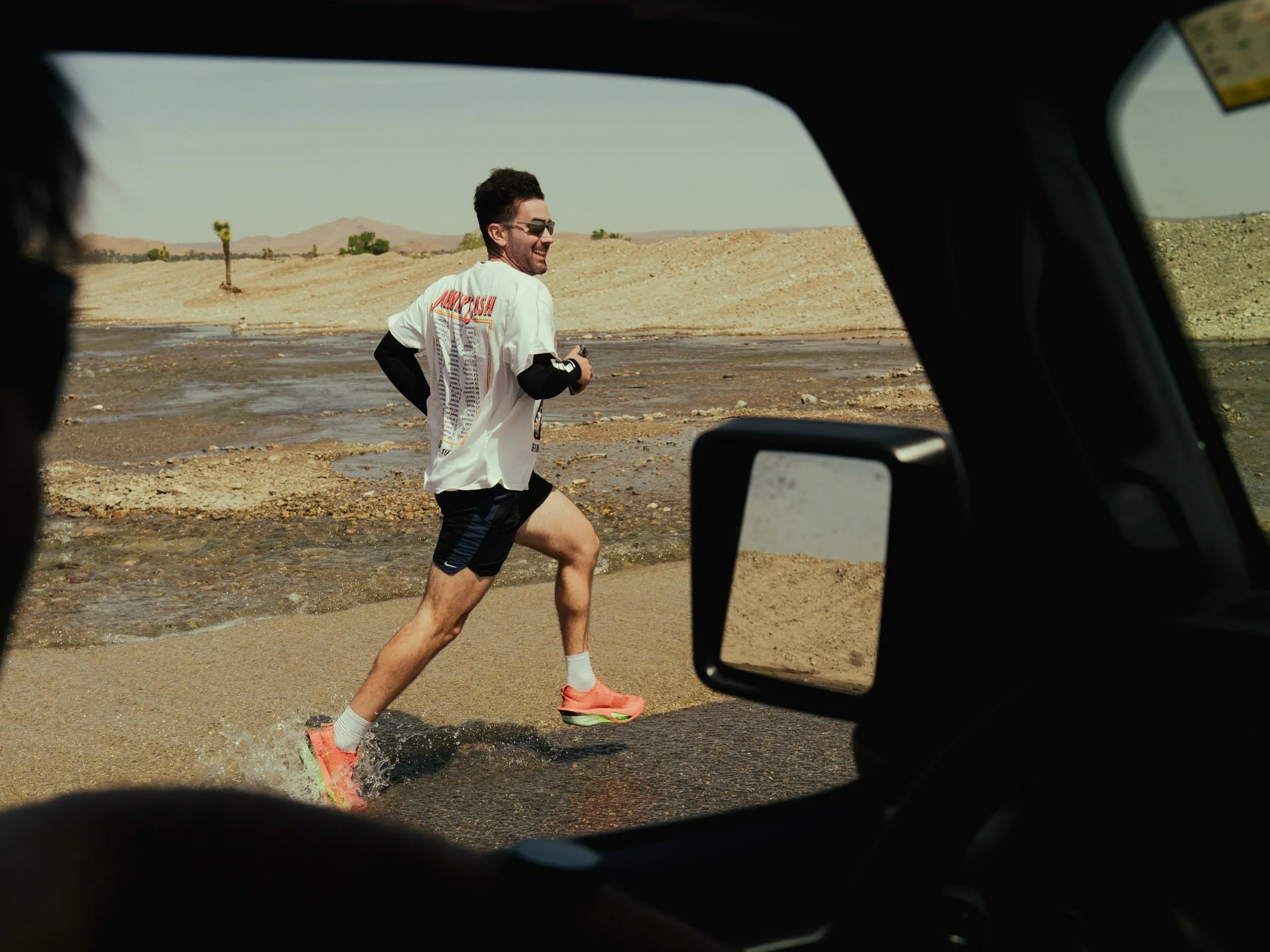 A man running in a desert area through shallow water, wearing sunglasses, a white t-shirt, black shorts, and pink running shoes, with a smile on his face.