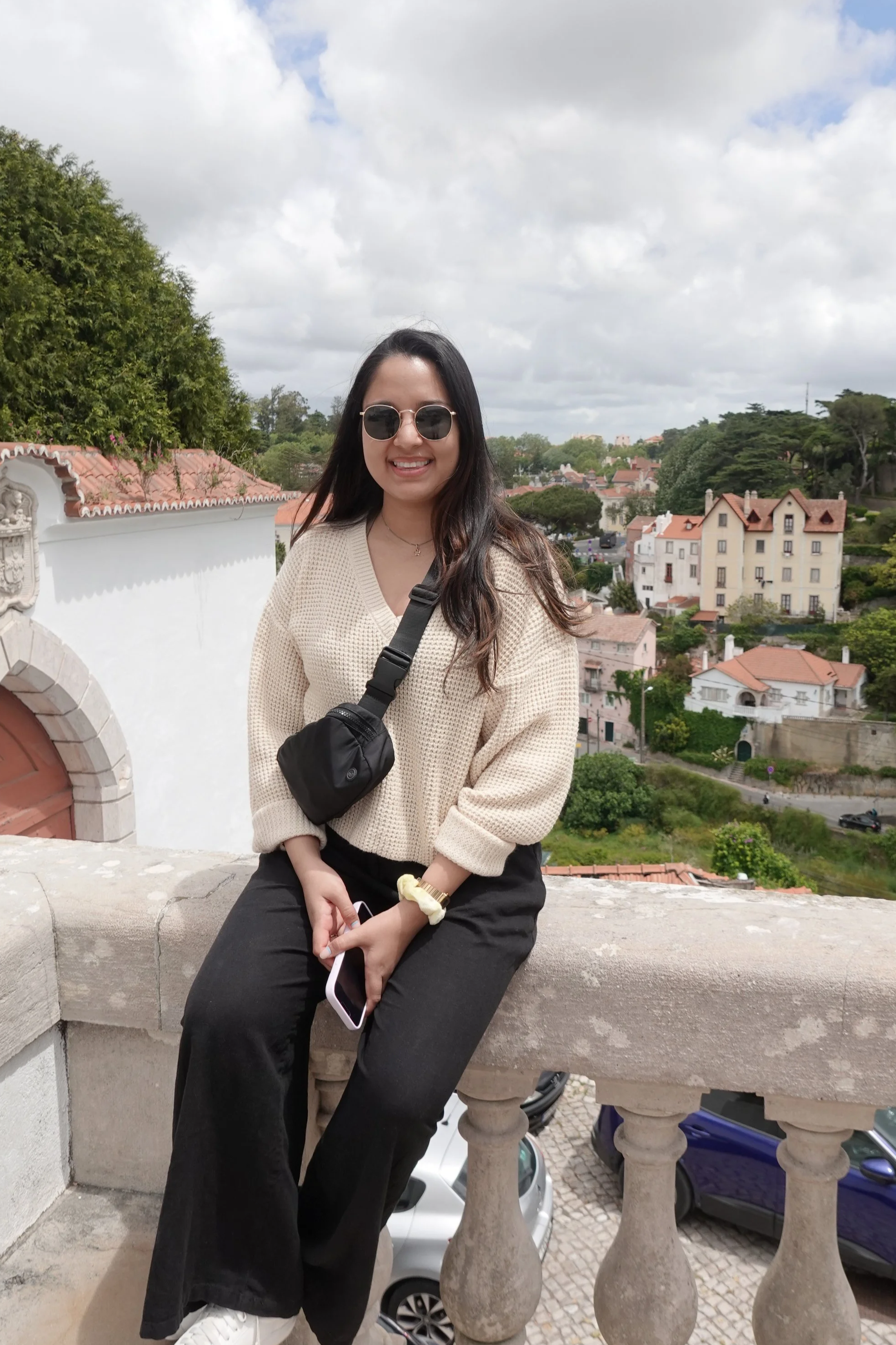 A young woman with long dark hair and sunglasses smiling while sitting on a stone ledge in front of a scenic view of houses and trees under a cloudy sky, wearing a beige sweater, black pants, and white sneakers, holding a smartphone.