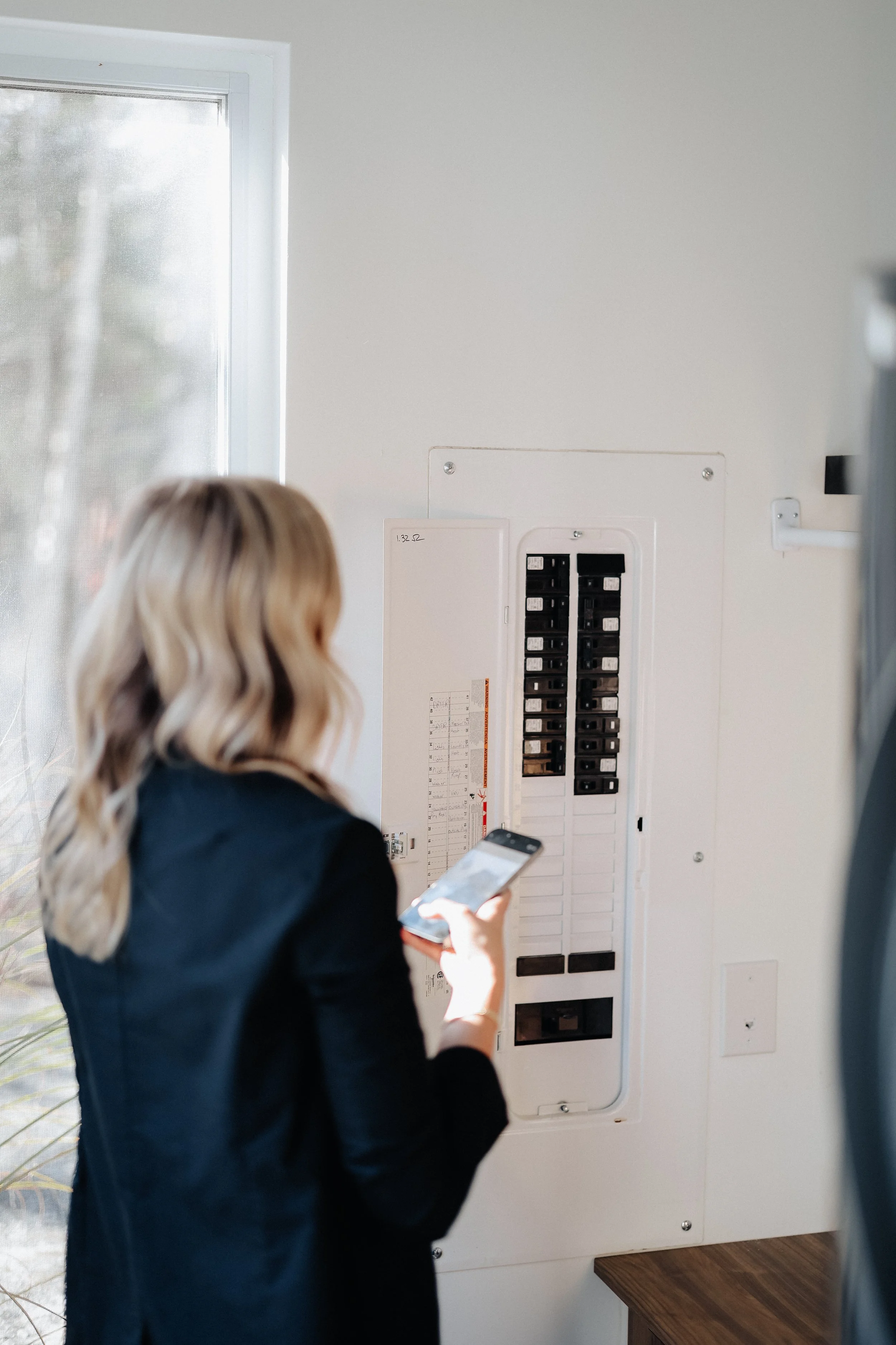 A woman with wavy blonde hair, wearing a black blazer, standing near an electrical panel on a white wall, looking at her smartphone.